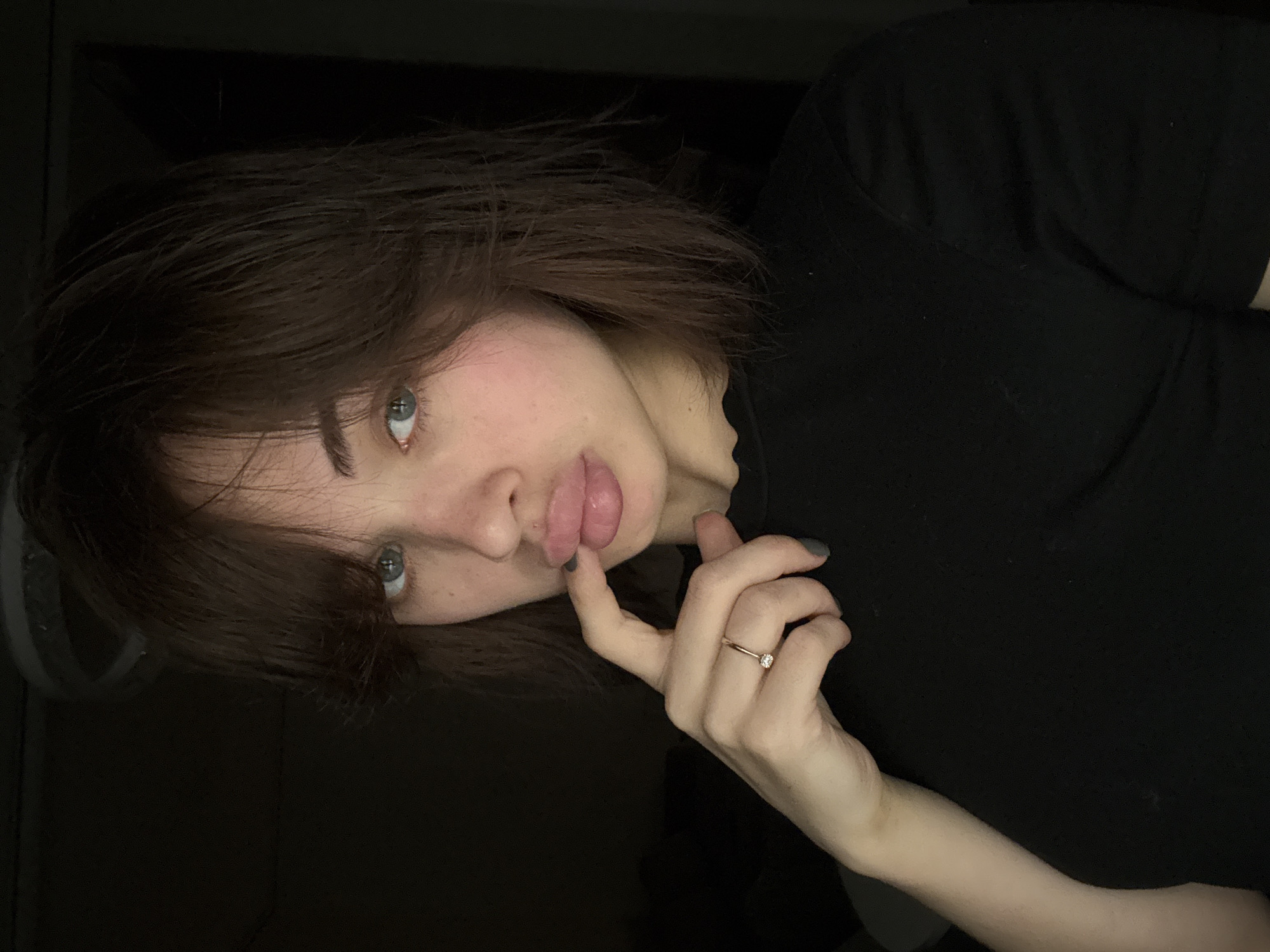 A young woman with shoulder-length brown hair and blue eyes is posed against a dark background. She is wearing a black top and a gold ring is visible on her left hand. She is holding her finger up to her lips, which are slightly parted. The lighting in the scene is dim. There is no information to confirm this photo was taken in Лобаново, Russia.