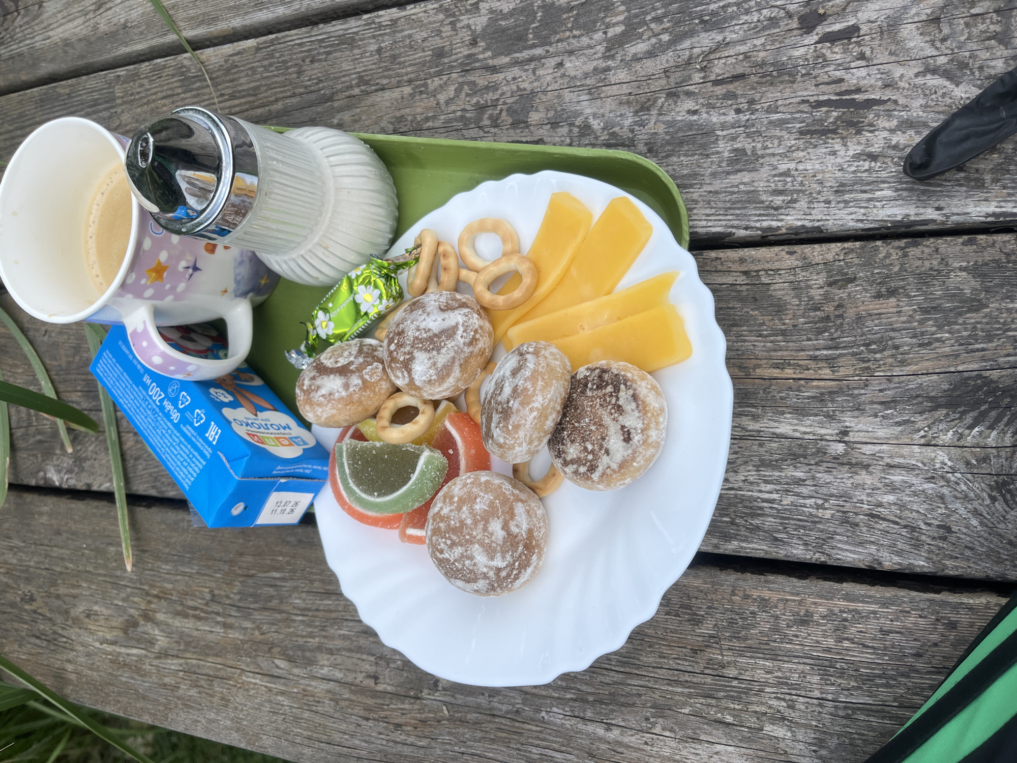 A high-angle, close-up shot captures a picnic scene on a weathered wooden table. A white plate, positioned on a green plastic tray, is laden with an assortment of food items. These include several round, powdered cookies, two slices of bright yellow cheese, three ring-shaped crackers, and two colorful fruit jelly slices, one green and one orange. Adjacent to the plate is a clear glass sugar dispenser filled with white sugar and a metallic top. A white ceramic mug, adorned with pink and purple stars and moon motifs, contains a beverage with a frothy top, likely coffee. A small blue carton, possibly of milk, rests behind the mug. The background features a blurred green plant, suggesting an outdoor setting. The lighting is natural and diffused, casting soft shadows.