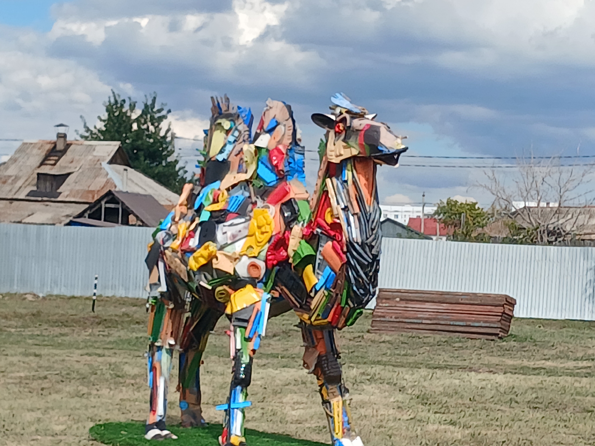 In Makinsk, Kazakhstan, a vibrant, life-sized sculpture of a camel, constructed entirely from colorful recycled materials, stands in an open grassy field under a cloudy sky. The sculpture is intricately assembled from a diverse array of discarded items, including plastic bottles, containers, and various scrap materials, forming its hump, body, legs, and head. The camel's head features red eyes and a blue object on its forehead. Behind the sculpture, a corrugated metal fence runs horizontally across the frame, with a pile of wooden planks stacked to its right. In the background, several low-rise buildings with rustic roofs are visible, and trees are scattered throughout the landscape. The ground around the sculpture is covered in dry grass, with a small patch of artificial green turf beneath the camel's feet. A striped post is positioned to the left of the sculpture.