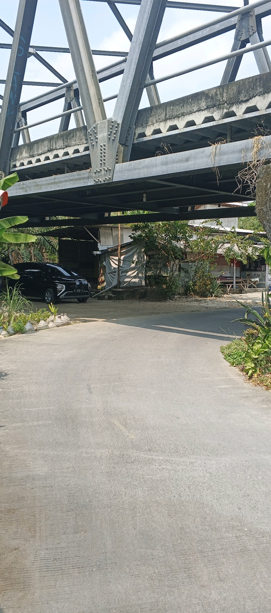This media file depicts a scene under a large, gray steel truss bridge. The bridge's structure, with its numerous bolts and beams, dominates the upper portion of the image. Below the bridge, a paved road curves towards the right, leading into a more populated area.
On the left side of the road, a black car is parked. Beyond the car, a small, rustic structure is visible, partly covered with a gray tarp. Various plants and greenery are scattered around the scene, with some banana leaves on the far left and lush vegetation on the right side of the road. The ground is paved with asphalt, showing some wear and tear.
The time of day appears to be daytime, with sunlight casting shadows, suggesting a clear or partly cloudy sky, though the sky itself is not prominently visible. The setting seems to be a rural or semi-urban area, possibly a roadside location in Laren, Indonesia, given the context. There are no people visible in the image.
A license plate on the black car is partially visible, showing "N 1920 F". There is also some faint graffiti or writing on the bridge's structure in the upper left corner, but it is illegible. The overall impression is one of an everyday, perhaps slightly overlooked, location.