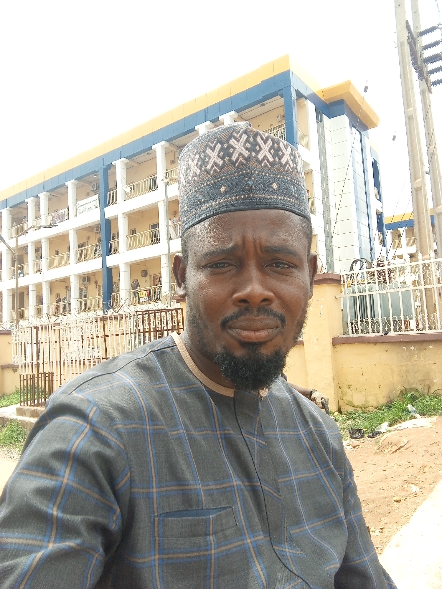 A man wearing a patterned cap and a grey and blue checkered shirt is standing in front of a multi-story building. The building has a blue and yellow facade with balconies on each floor. There are electrical poles and wires visible to the right of the building. The ground in front of the building is a dirt path with some scattered debris. The scene appears to be in an urban environment, likely Abuja, Nigeria, given the architectural style and presence of electrical infrastructure typical of the region.