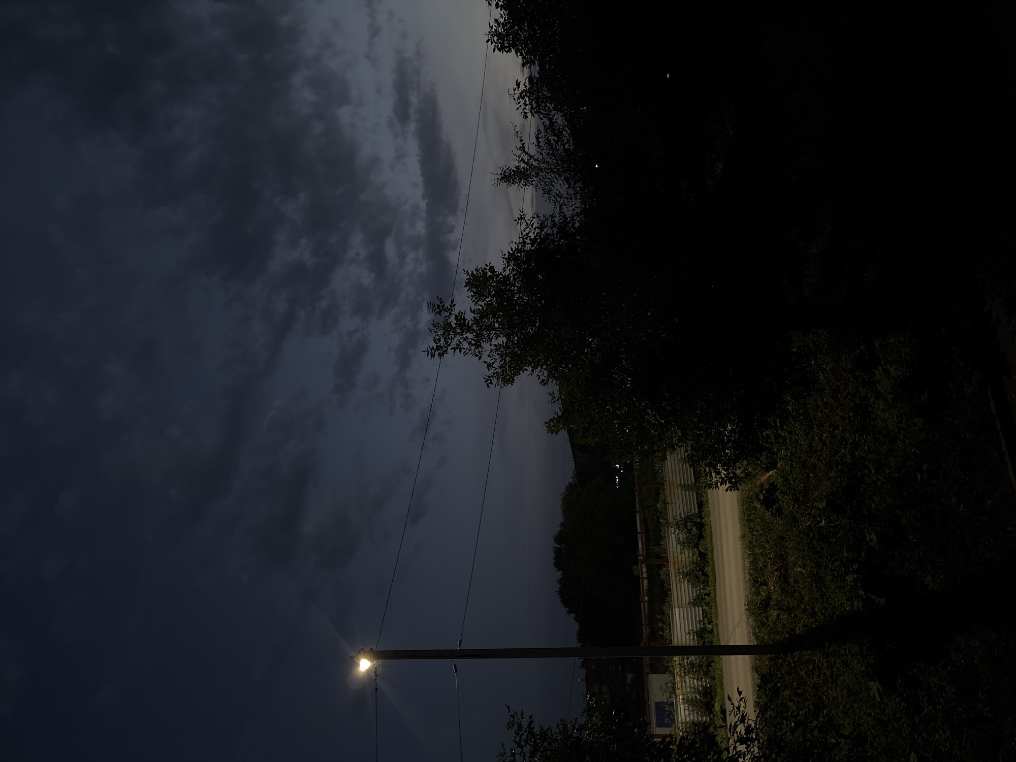This media file depicts a street scene at dusk or dawn in Inskoy, Russia. The sky is filled with dark, dramatic clouds, suggesting an overcast or stormy weather condition. A single streetlight illuminates the foreground, casting a warm glow against the dark sky. Silhouetted trees and foliage frame the scene on the right and in the bottom corners.
A gravel road is visible, stretching into the distance alongside a corrugated metal fence. The lighting conditions are low, with the overall impression being somber and slightly mysterious. The image captures a moment of transition between day and night, marked by the artificial light of the streetlamp and the natural dimness of the twilight sky. There is no visible text in the media file.