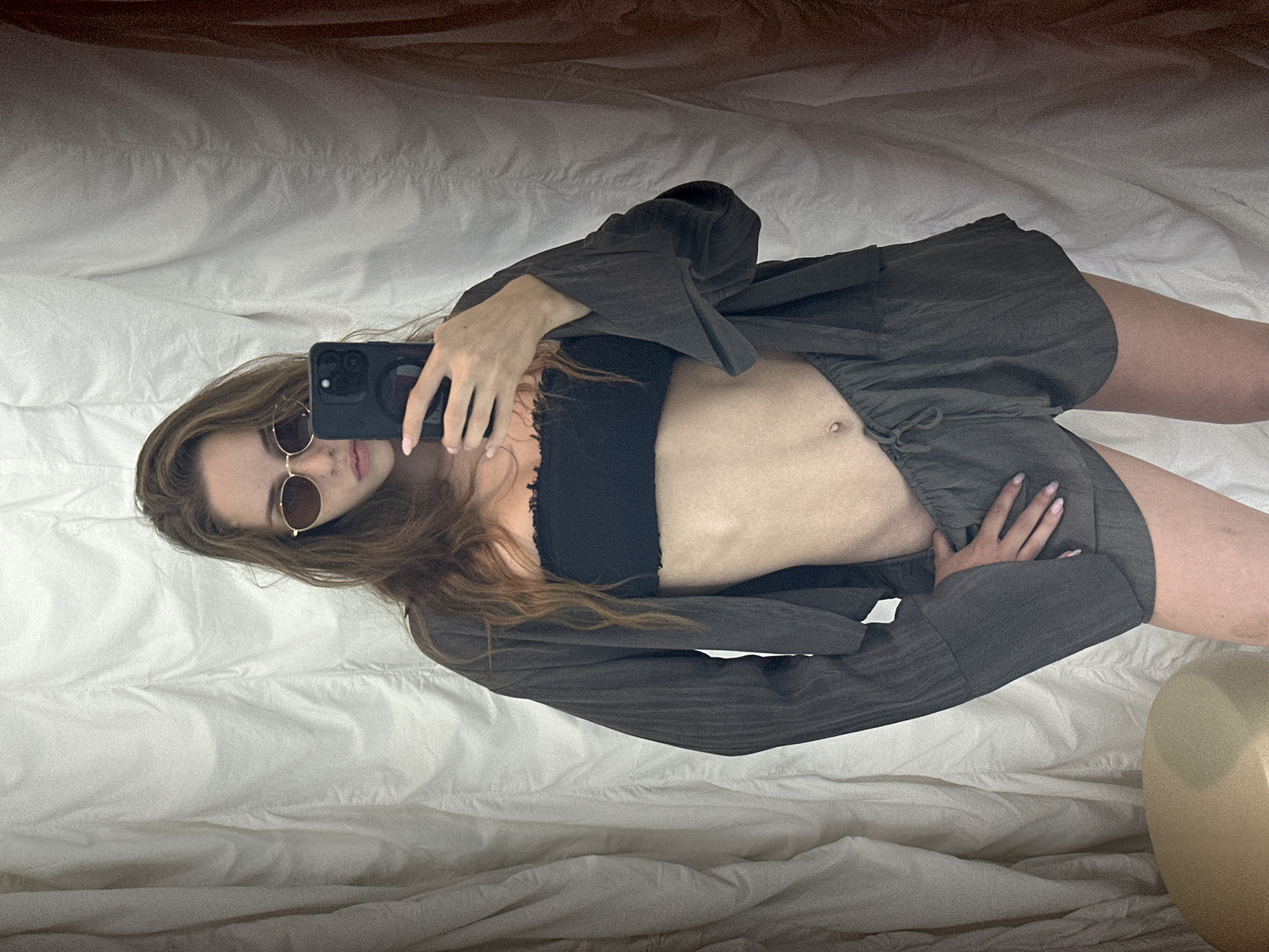 A female individual stands in an indoor setting in North Kuta, Indonesia, capturing a mirror selfie with a black smartphone held in her right hand. She possesses long, wavy brown hair and wears oval-shaped, gold-rimmed sunglasses. Her attire consists of a black strapless bandeau top with a visible raw-cut or fringed upper edge, exposing her midriff which features a small navel piercing. She is additionally dressed in a dark olive-grey, long-sleeved, open-front jacket or cardigan with a textured fabric, and matching drawstring shorts. Her fingernails are manicured with a light pink polish. Her left hand rests on her left hip, partially obscured by the shorts. The background behind her is characterized by light-colored, vertically pleated or draped fabric, possibly curtains or a textured wall, with a darker vertical element on the right. A rounded, light-toned object is visible in the lower left corner of the frame.