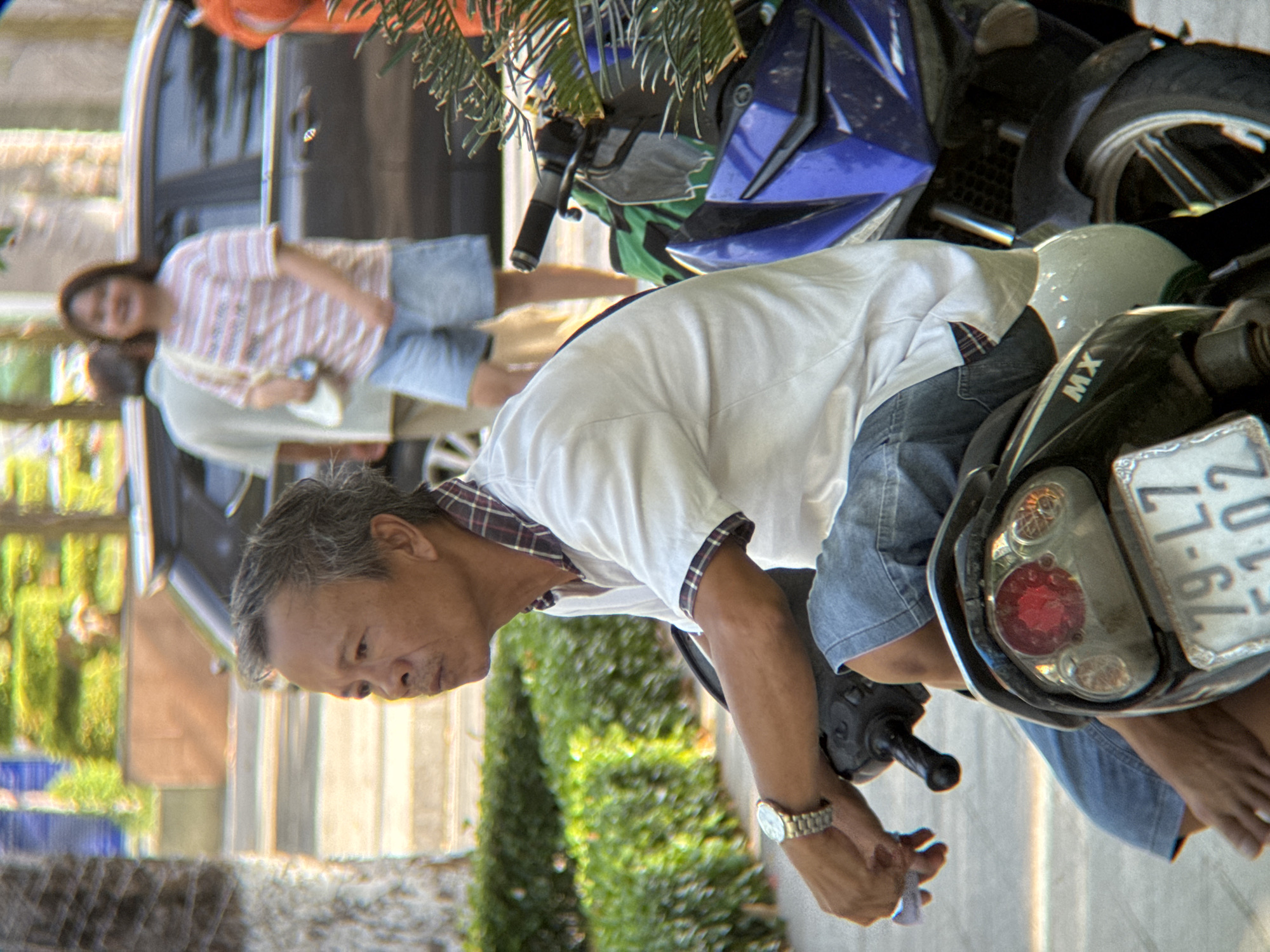 The image depicts a man sitting on a motorcycle in what appears to be a street scene in Nha Trang, Vietnam. The man, who has graying hair and is wearing a white short-sleeved collared shirt and denim shorts, is looking to his left. A silver watch is visible on his left wrist. The motorcycle is black and blue with visible "MX" branding. A license plate with the characters "79-L7 51.02" is attached to the rear of the motorcycle. In the background, a woman in a striped t-shirt and denim shorts is walking, and a person in a light-colored shirt is also visible. The environment includes a blurred background of buildings and greenery, suggesting an urban setting. The lighting indicates it is daytime.