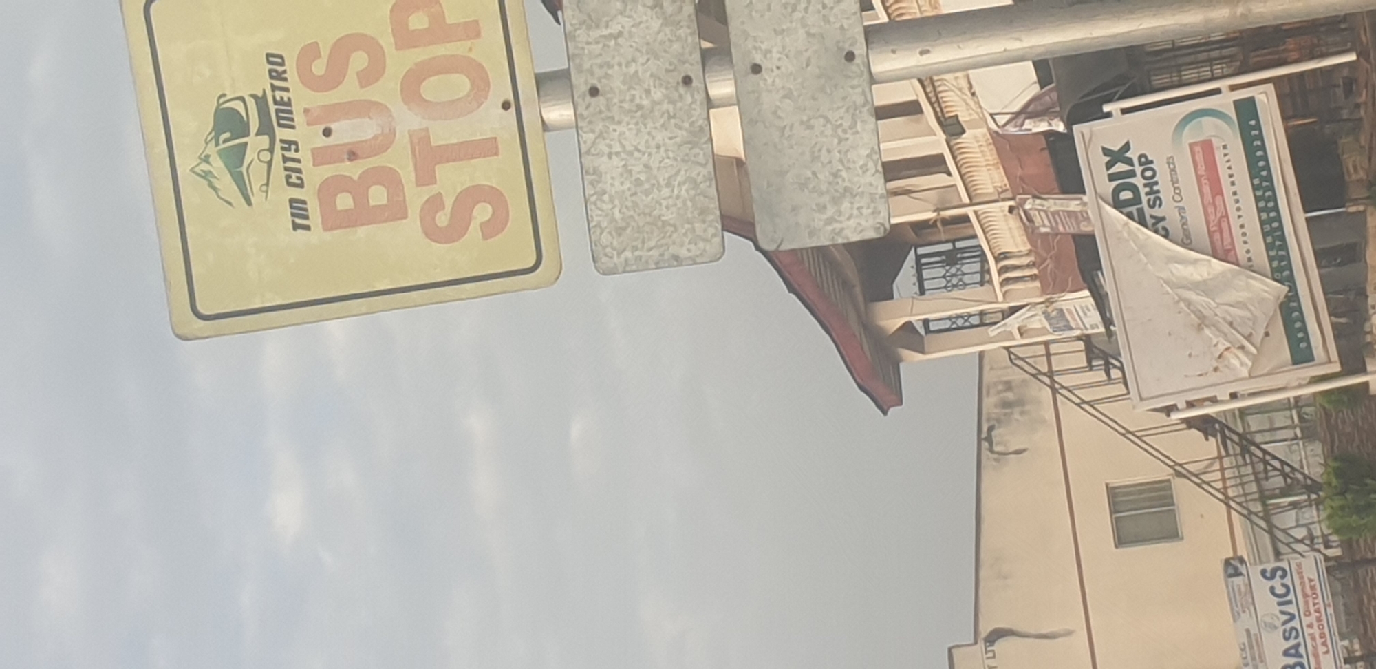 The image depicts an outdoor scene with a clear sky, suggestive of daylight. In the foreground, a yellow sign with black and orange lettering indicates "TIN CITY METRO BUS STOP." Below this sign, two metallic rectangular plates are attached to a pole.
In the background, a building with a red roof and beige walls is visible. It features a balcony with white railings and metal grilles on the windows. An external staircase leads up to the building. Several signs are attached to the building and stand in front of it. One sign is partially obscured by a torn white sheet and advertises "EDIX CY SHOP General Contracts." Another sign, partially visible on the left, reads "BASVICS Medical & Diagnostic LABORATORY." The overall environment appears to be an urban setting, possibly a roadside, with the presence of commercial establishments and public transportation infrastructure. The phrase "Tin City" is a known nickname for Jos, Nigeria, suggesting the location.