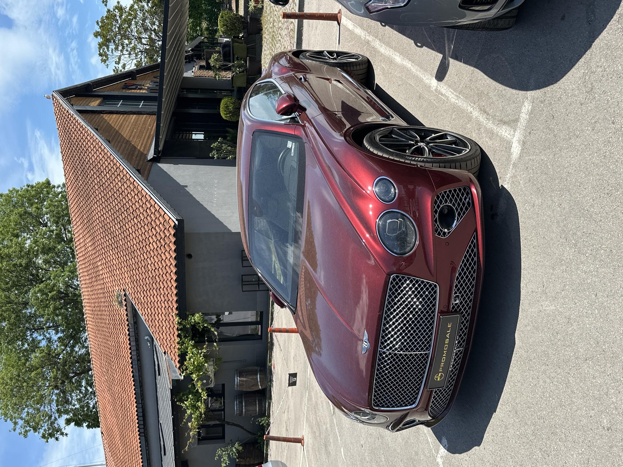 A maroon Bentley Continental GT is parked in an outdoor parking area next to a building with a steeply pitched red tile roof. The car is facing forward, and part of another vehicle is visible parked next to it. The parking area appears to be paved with concrete. The location is Varna, Bulgaria.