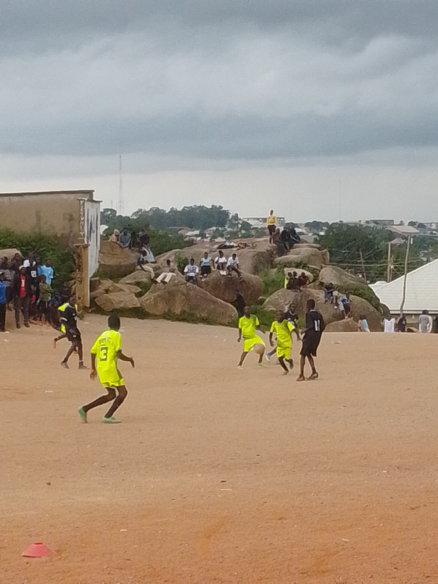 The image depicts an outdoor scene in an urban environment, likely a dusty or unpaved area, with a group of people engaged in a soccer match. The sky is overcast and appears cloudy. In the background, there are buildings, some scattered on a rocky hillside, and a tall communication tower.
In the foreground, several young men are playing soccer. One player in a bright yellow jersey with the number "3" on the back is in motion, seemingly dribbling or about to kick the ball. Another player in a similar yellow jersey is nearby. A player in a black jersey with the number "11" is also visible. Several other individuals, likely spectators or teammates, are gathered on the side of the playing area and on the rocky formation in the mid-ground. One person in a yellow shirt is standing on top of the rocks in the background. The ground is predominantly sandy or dusty brown. A red cone is visible in the lower left corner of the image. The overall impression is of an informal soccer game taking place in a community setting.