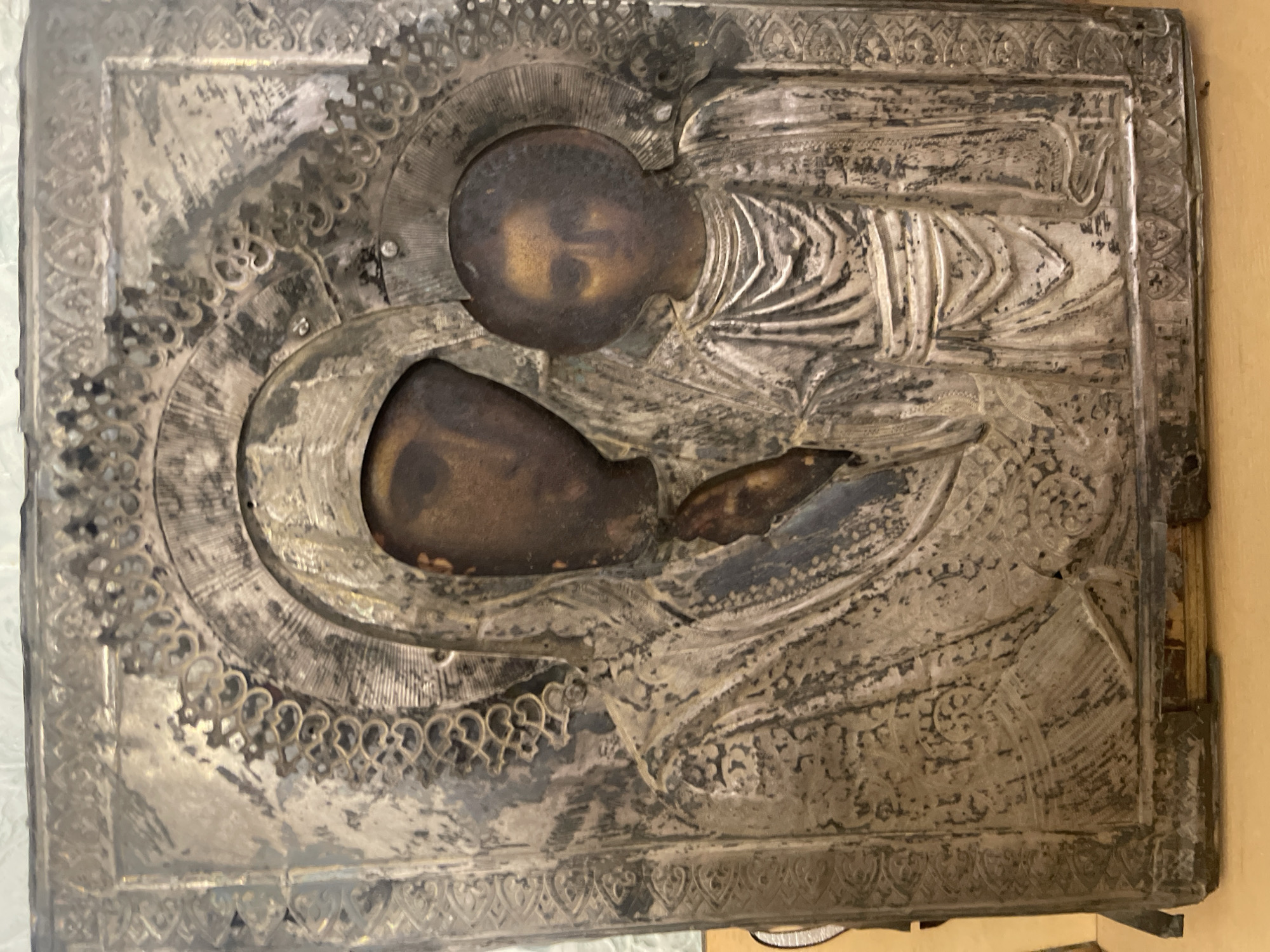 The image shows an aged, silver-plated icon depicting the Virgin Mary holding the infant Jesus. The icon is rectangular and appears to be made of metal, likely silver or silver-plated. The raised, embossed figures of Mary and Jesus are the focal point, with their faces appearing worn and tarnished. The surrounding silver surface exhibits detailed patterns and designs, including floral motifs and what looks like draping fabric. The icon shows signs of significant wear and tarnishing throughout. There is no specific location associated with the icon based on this image. The city and specific details regarding its origin are unknown.