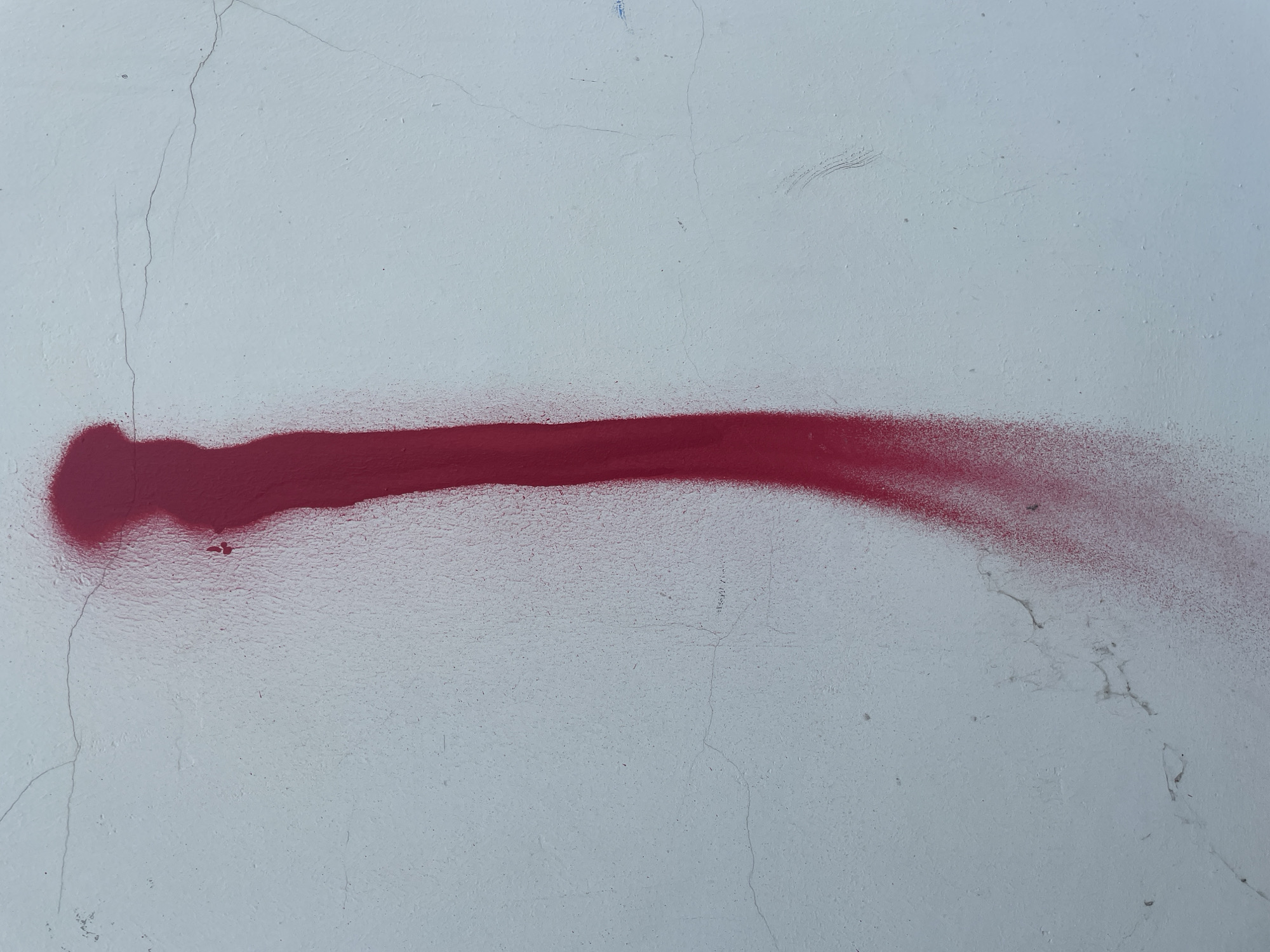 An abstract, vertical streak of red spray paint adorns a pale, textured wall in an outdoor setting. The paint application is uneven, with a broader, more opaque section at the top that tapers into a finer, diffused spray towards the bottom. The wall exhibits subtle cracks and a slightly uneven surface, indicative of age or weathering. The lighting suggests an overcast or diffused daylight condition. There are no people or other discernible objects in the frame.