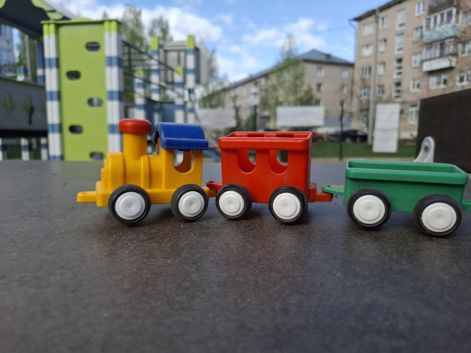 A toy train sits on a grey surface, likely a picnic table, outdoors. The train consists of a yellow locomotive with red and blue accents, a red carriage, and a green carriage. Behind the train, there is playground equipment, with a green and white structure prominently visible. Apartment buildings are visible in the background, suggesting an urban area. The sky is blue with white clouds. The location is in the city of Chaikovsky, Russia.
