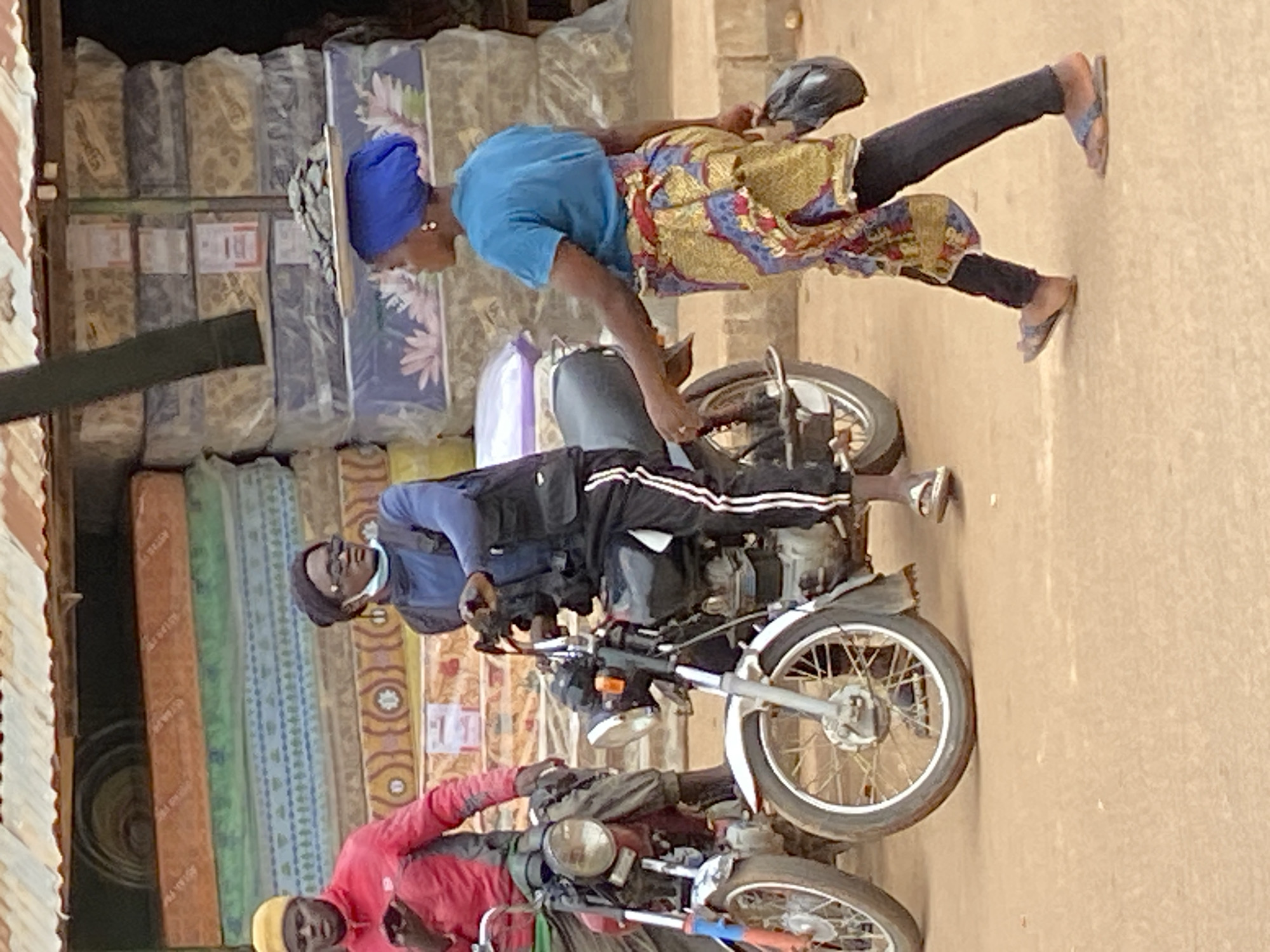 Two men on motorcycles and a woman walking are in a market setting in Bukuru, Nigeria. The man on the left, wearing a red jacket and a yellow beanie, is on a red motorcycle. The man on the right, wearing a black beanie, sunglasses, a blue jacket over a vest, and black pants with white stripes, is on a silver motorcycle. A woman in a blue headscarf, blue t-shirt, and a colorful skirt is walking to the right, carrying a black bag in her right hand and a tray with items on her head. Behind them, stacks of mattresses are visible, some wrapped in plastic. The ground is dusty and brown.