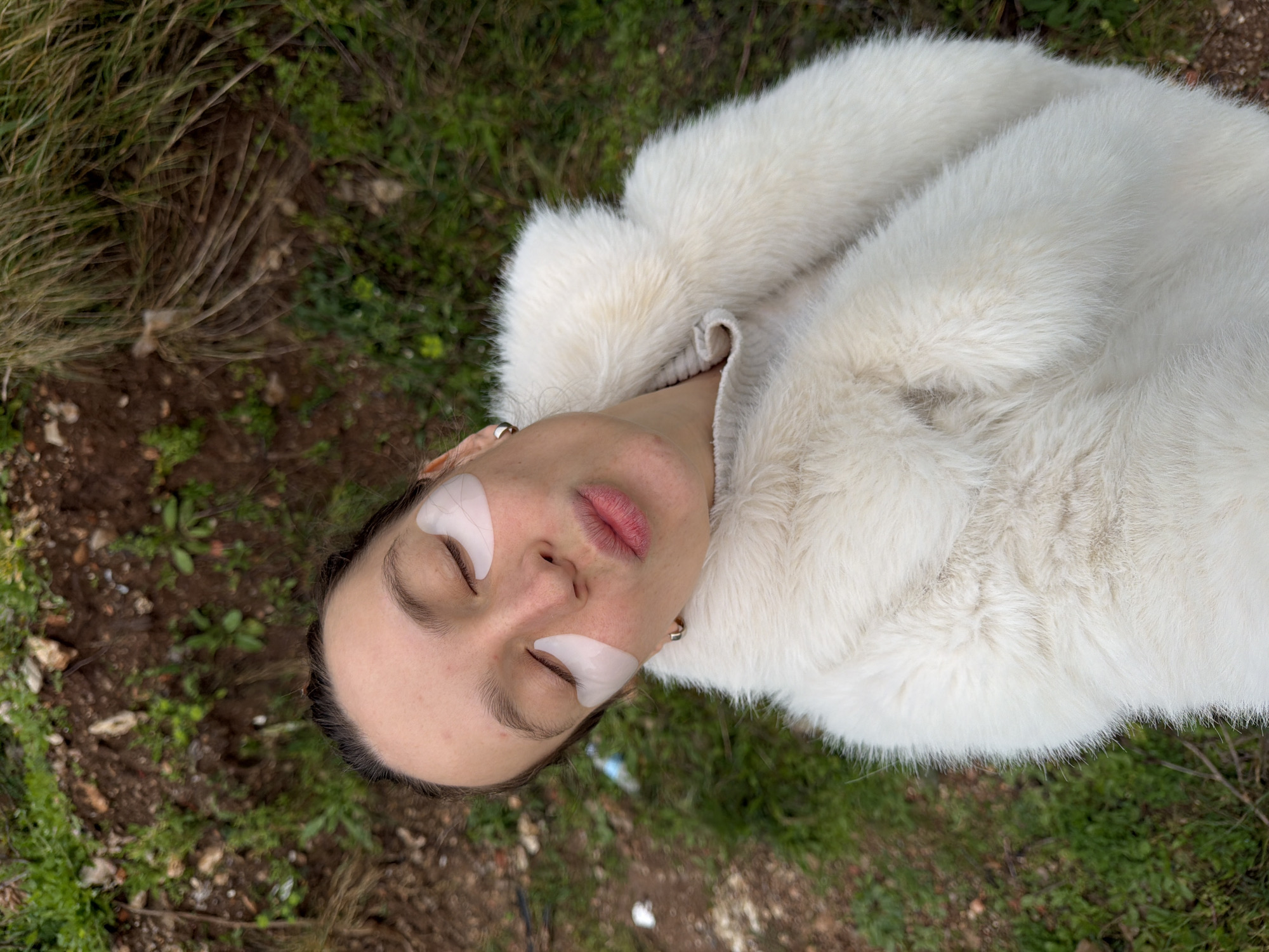 The image depicts a single individual positioned outdoors in a natural, uncultivated area, consistent with a hillside or undeveloped plot within or on the periphery of Budva, Montenegro. The subject, appearing to be female, is captured from an overhead perspective, looking upwards towards the camera. Her eyes are closed, and her lips are pursed. Two light-colored, translucent eye patches are applied beneath each eye. A small, silver-toned hoop earring is visible on her right earlobe. She is dressed in a thick, white or off-white faux fur coat, with a light-colored, ribbed sweater visible at the neckline. The background consists of uneven ground, primarily brown soil interspersed with various green vegetation, including sparse grass and small plants, along with some dry, yellowish-brown brush. No specific city landmarks are visible in the natural environment.