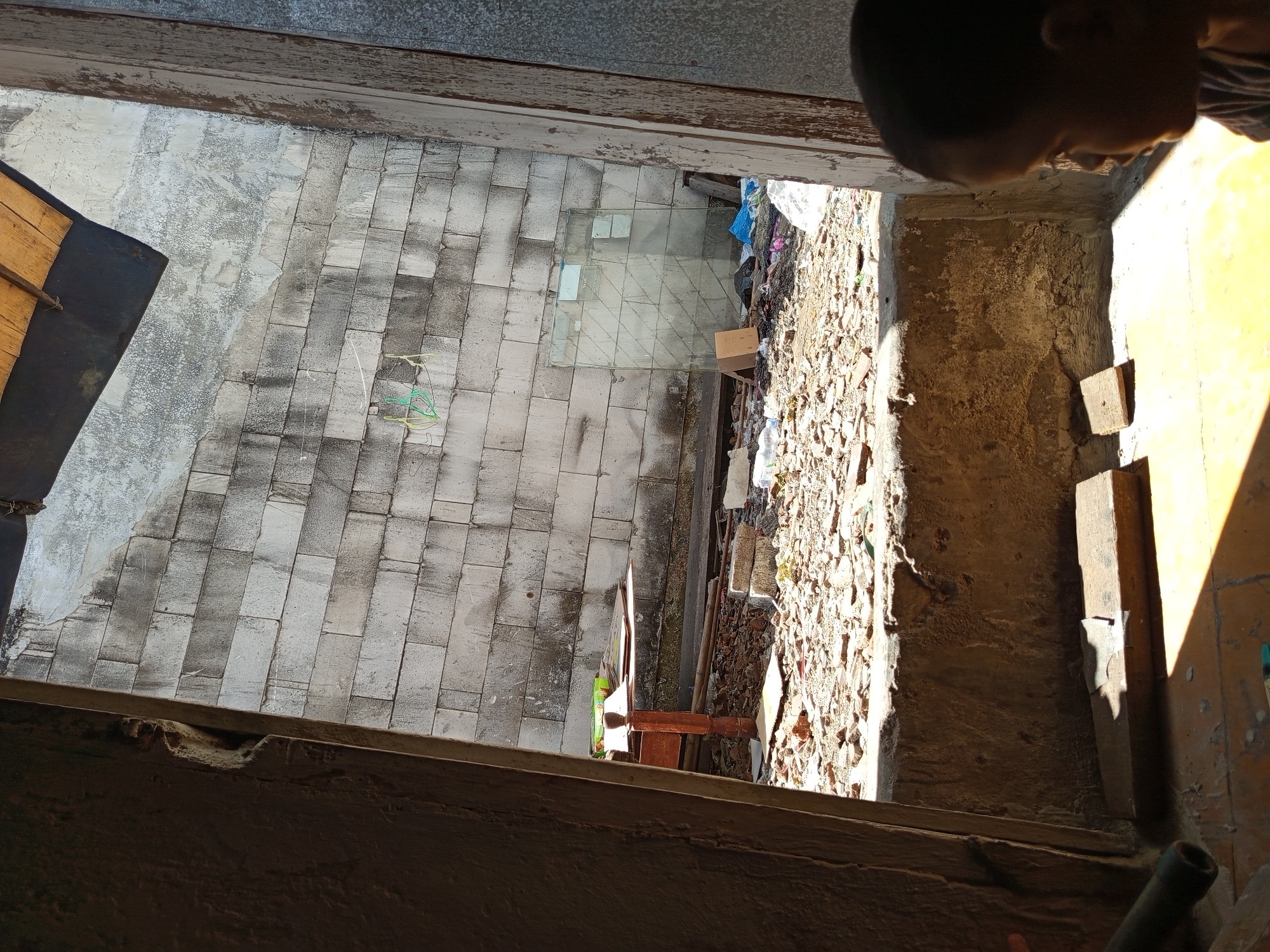 The image shows a young child in the foreground looking out from an opening. The opening frames a scene of a yard filled with debris, including broken bricks, trash, and a piece of glass. A wooden table with some items on it is visible against a wall constructed of cinder blocks. The setting appears to be an outdoor space in Tebel, Indonesia, characterized by the sunlight and the materials of the surrounding structures.