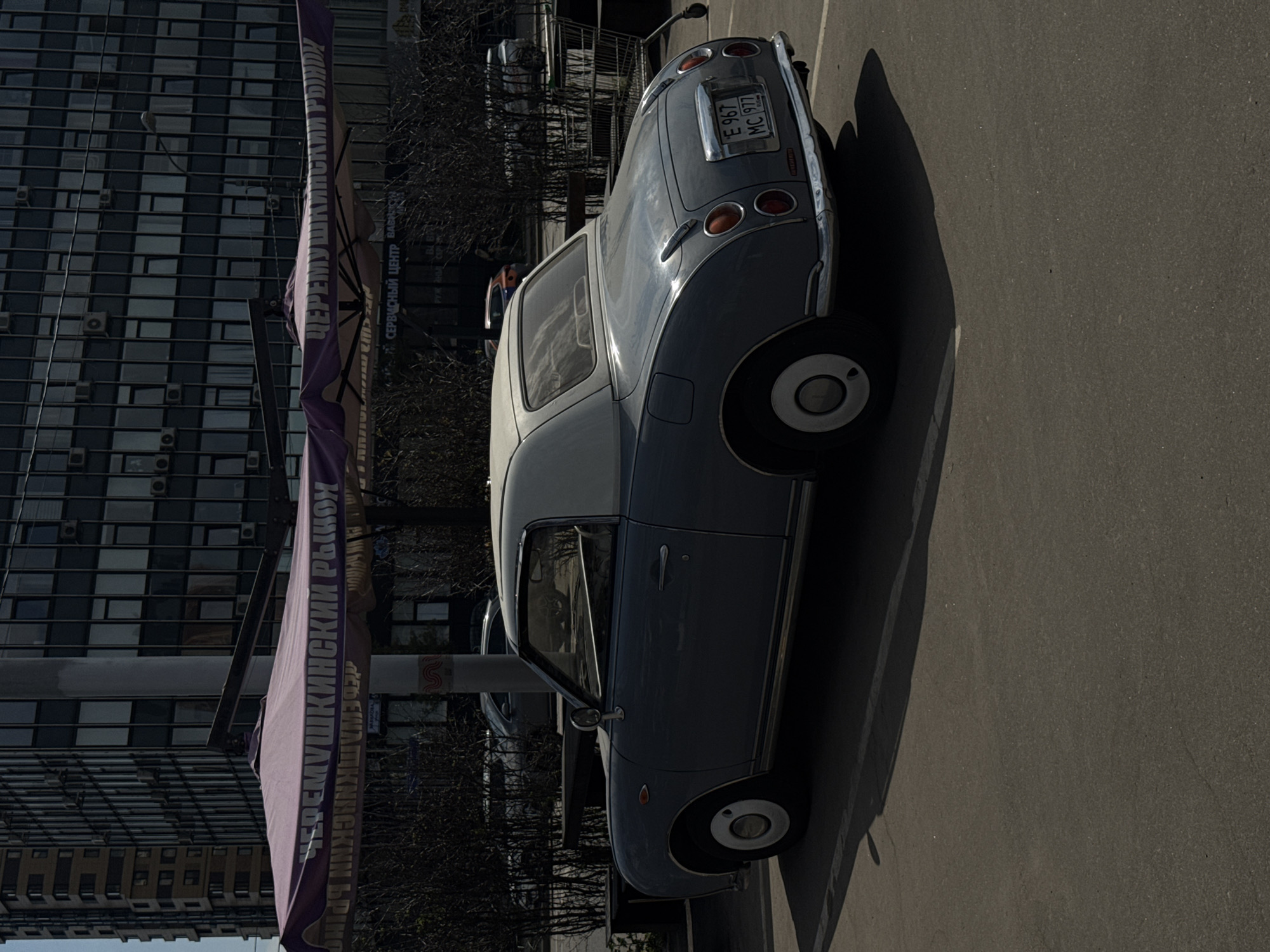 The image shows a parked, light blue and white, retro-style car in Moscow, Russia. The license plate reads "E 967 MC 977." The car is parked on asphalt in front of a building. A large banner on a pole with Cyrillic text, "ЧЕРЕМУШКИНСКИЙ РЫНОК" ("Cheremushkinsky Market"), is visible next to the car. A modern building with many windows stands in the background.