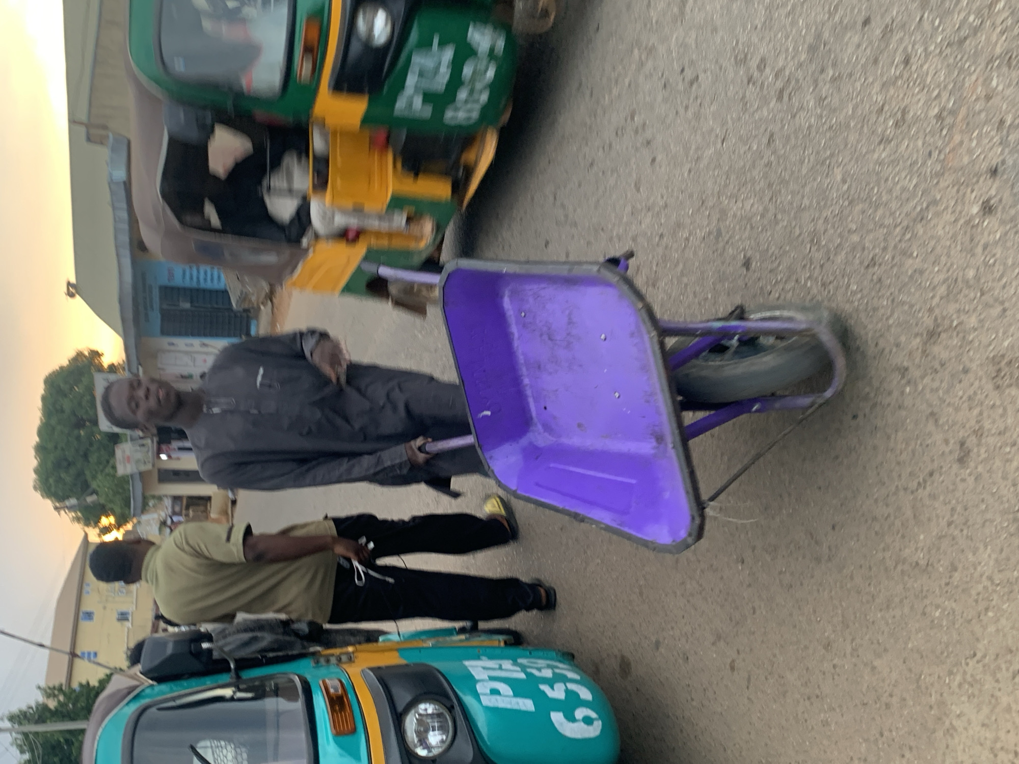 In Jos, Nigeria, at dusk, a young man in a dark gray tunic and trousers pushes a vibrant purple wheelbarrow down a paved street. Behind him, another man in a light olive green polo shirt and black pants walks away from the camera, holding a white object in his right hand. To the left of the frame, a teal and yellow auto-rickshaw, possibly a "Keke Napep," is parked. It has white lettering on its side reading "PTA" and "6559". To the right, another auto-rickshaw, predominantly green and yellow, is partially visible, with similar white lettering that appears to read "PTA" and "8034". In the background, buildings with yellowish walls and tiled roofs are visible, suggesting a residential or commercial area. The sky is a warm orange and yellow, indicating the setting sun. The overall scene depicts a common street view in Jos, likely during the evening commute or a period of commercial activity.