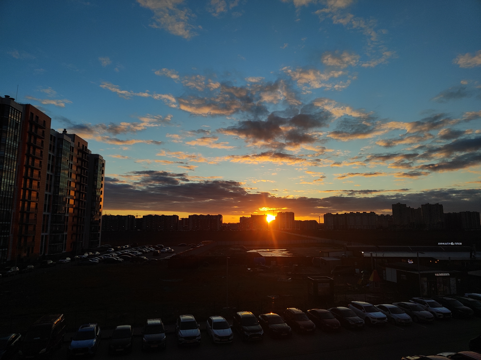 The image presents an urban landscape at sunset, characterized by a striking interplay of light and shadow. The upper half of the frame features a clear blue sky interspersed with scattered clouds. Many of these clouds are illuminated from below by the setting sun, displaying warm orange, pink, and golden hues, while others appear darker gray and purple.
The sun itself is a bright, intense orb positioned low on the horizon, creating a prominent starburst effect and casting a strong glow that silhouettes the cityscape. On the far left, a tall, modern apartment building stands, its numerous windows and multi-toned facade catching and reflecting the last light of the day. Along the distant horizon, a dense line of other high-rise buildings forms a dark silhouette against the fiery glow of the sunset.
The mid-ground and foreground are largely in shadow, revealing extensive parking lots filled with numerous parked cars, mostly dark, with some reflecting subtle glints of light. Patches of dark ground, possibly grass or undeveloped land, are also visible, along with some smaller, low-rise structures. A wet area or puddle can be seen in the mid-ground.
No people are visible in the scene. The overall impression is one of a peaceful urban evening. Visible text on a sign in the mid-ground (discernible in provided crops) reads "ХИНКАЛИ & ПЛОВ" and "WILDBERRIES".