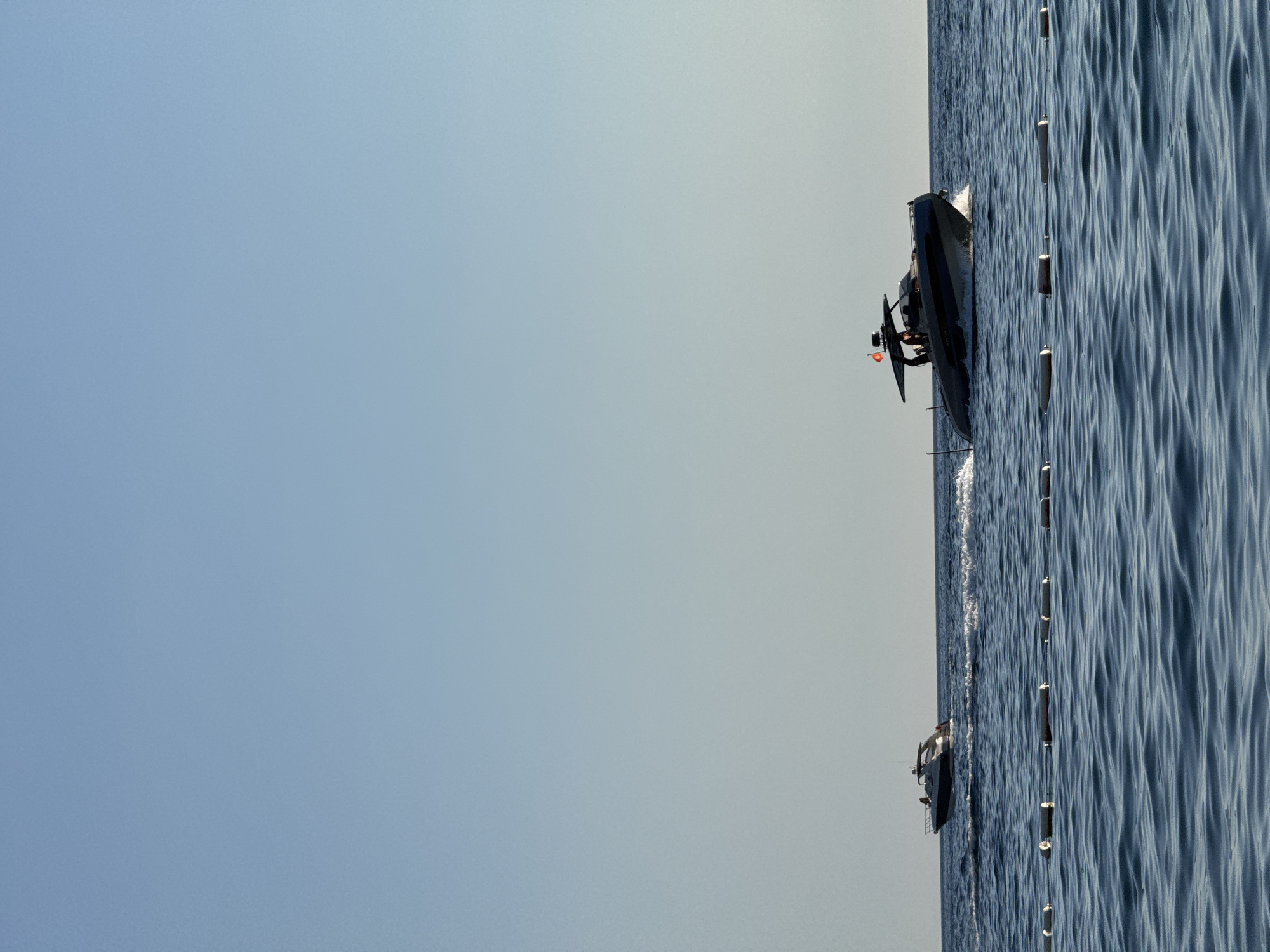 The image shows a body of water with two boats moving across it under a clear blue sky. In the foreground, a line of white buoys with red and white stripes floats on the surface of the blue water, marking a boundary. The water is choppy with visible waves.
The larger boat, positioned in the mid-right of the frame, is a sleek, modern speed boat. It is dark gray with blue accents and has several people on board, visible in silhouette. The boat is creating a white wake as it moves at speed. A small orange flag is visible at the stern of this boat.
A second, smaller white boat is visible in the mid-left of the frame, further away from the viewer. This boat also appears to be moving, creating a smaller wake.
The environment is consistent with a coastal area, potentially the Adriatic Sea near Sveti Stefan, Montenegro, given the clear skies and the presence of boats and buoys often found in tourist or recreational areas. The lighting suggests it is daytime, likely during the afternoon given the angle of the sun.