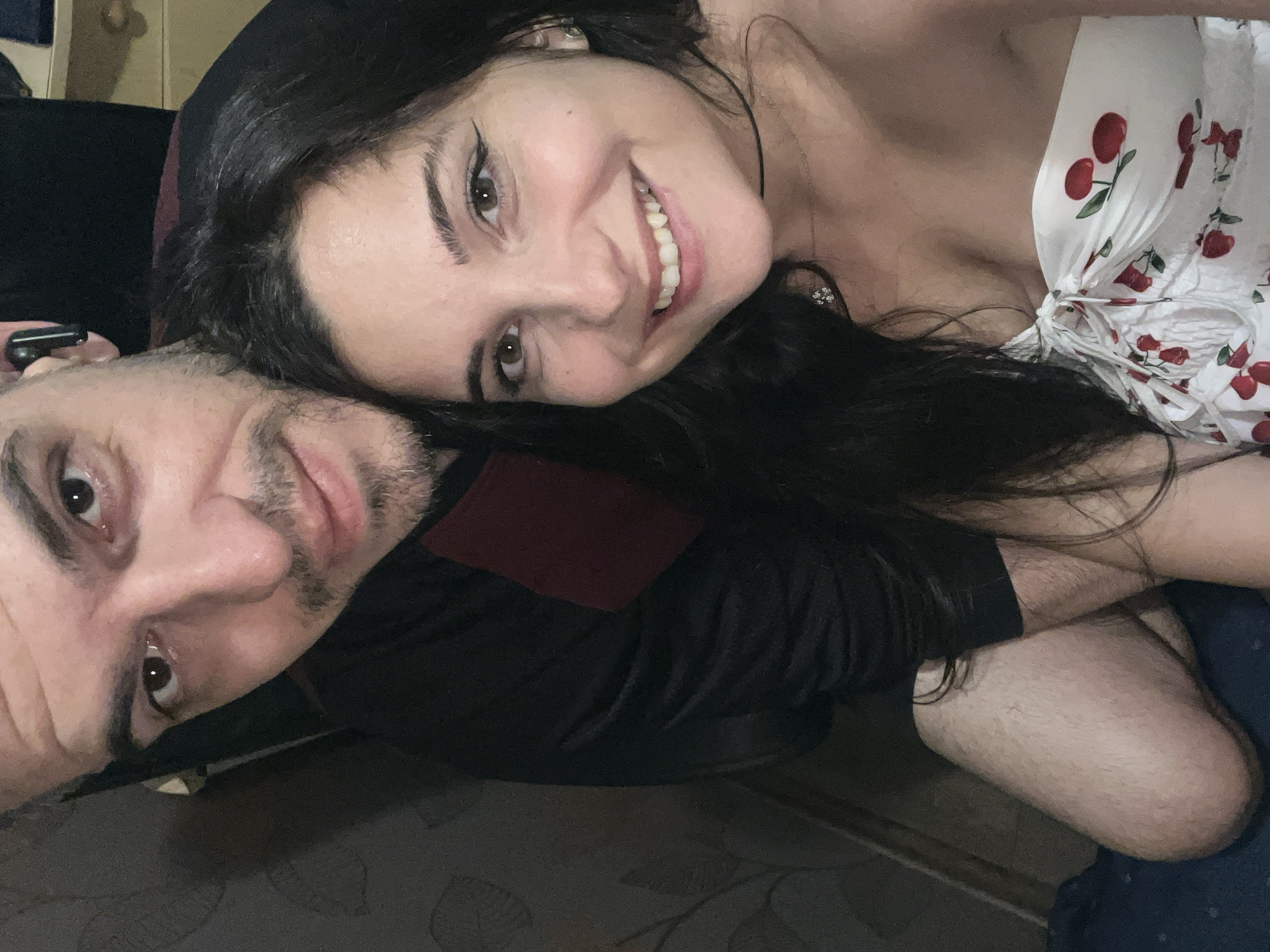 In an indoor setting, a man and a woman are captured in a close-up selfie. The man, on the left, has short, dark hair, a light beard, and is wearing a dark shirt with a maroon accent. He has an earbud in his left ear. The woman, on the right, has long, dark hair and is smiling. She is wearing a white strapless top with a cherry pattern and a delicate necklace. They appear to be in a casual, intimate pose. The background is dimly lit and shows a textured wall with wallpaper and hints of furniture. The location is specified as Sofia, Bulgaria, though no specific landmarks or identifying features of the city are visible in the image.