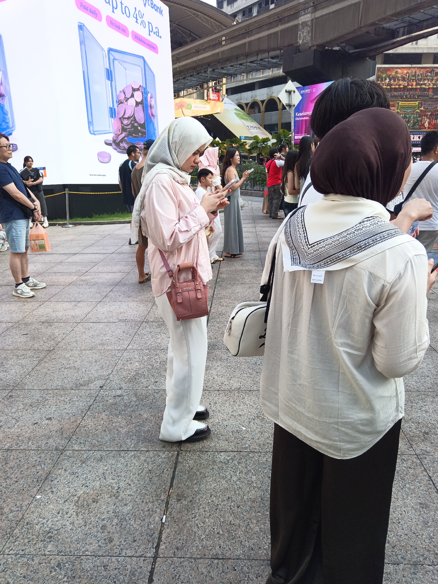 The image depicts a street scene in Kuala Lumpur, Malaysia, likely near a public transportation hub given the elevated structure visible in the background. A large billboard advertising a bank's offer of "up to 4% p.a." is prominent on the left. Several individuals are present on a paved pedestrian area. In the foreground, a person wearing a light pink hijab, a pale pink long-sleeved shirt, and white pants is looking at their phone. They are carrying a small, reddish-brown handbag. To their right, another person, partially obscured, wears a dark brown hijab, a cream-colored long-sleeved shirt with a patterned scarf around their neck, and dark pants. They are also looking at a phone. Other people, including a man in a blue shirt and shorts holding a plastic bag, a woman in a grey dress, and a person in a red shirt, are visible in the midground, some appearing to be walking or waiting. The environment suggests an urban setting with a mix of modern infrastructure and pedestrian activity.