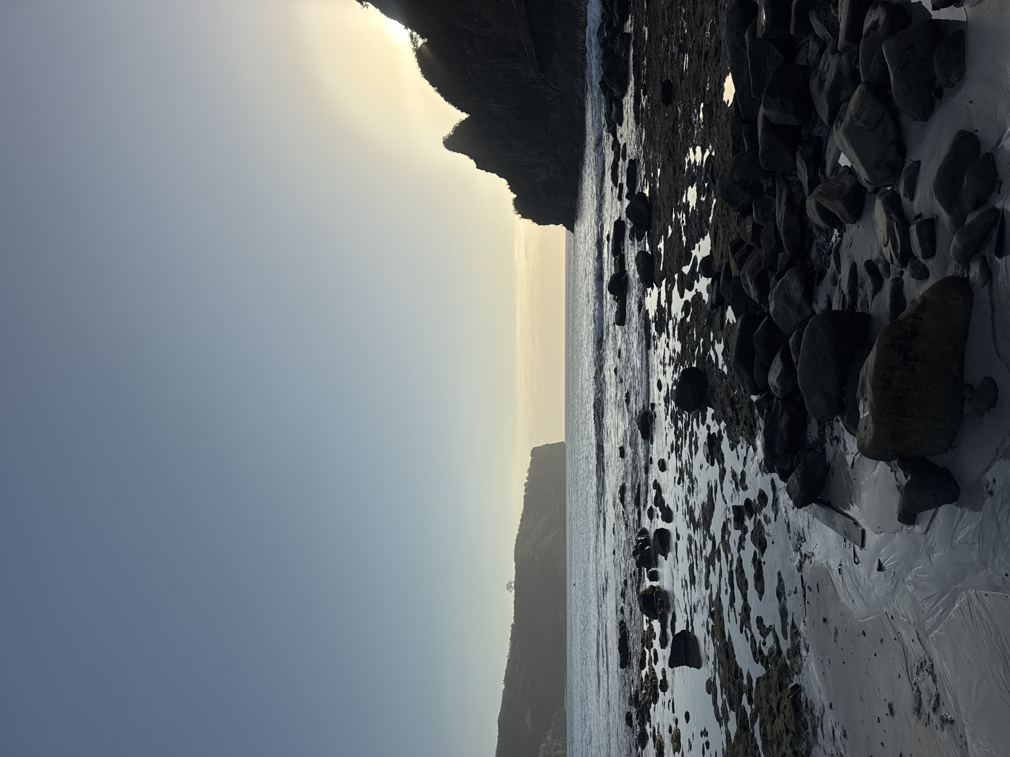 This is a vertical, eye-level, full shot of a rocky beach in Sekongkang, Indonesia, during what appears to be sunrise or sunset. The sky is clear and blue, fading to yellow near the horizon where the sun is partially obscured by a dark, silhouetted cliff face on the right. The ocean is calm, with small waves gently lapping the shore. The foreground is dominated by dark, wet rocks and patches of sand, some of which are reflecting the sky and light. In the distance, a large, tree-covered cliff juts out into the sea on the left, also silhouetted against the bright sky. There are no people or animals visible in the frame. The overall mood is serene and natural.