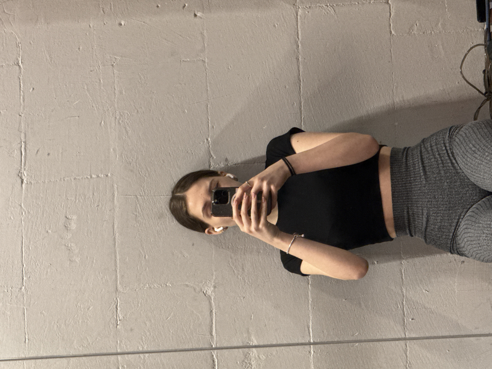 A woman is taking a selfie. She is wearing a black short-sleeved top and gray leggings. She is holding a smartphone in front of her face. She is wearing ear buds. The background is a gray wall that appears to be concrete blocks. The location is indeterminable, and there is no information visible in the image that can identify it as being in Sofia, Bulgaria.