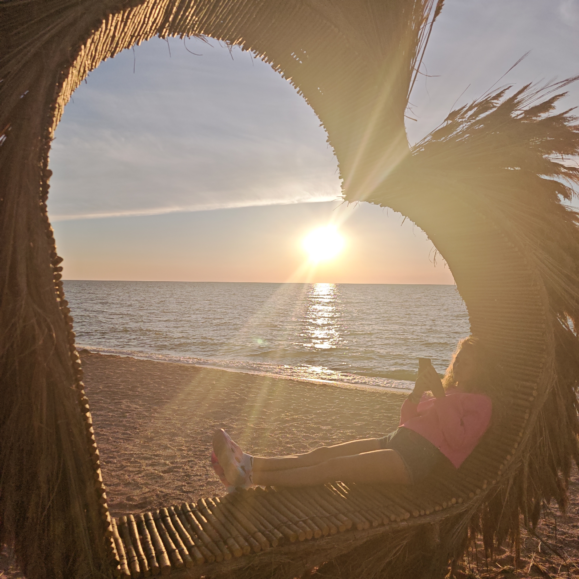 The image depicts a person sitting on a bench made of bamboo sticks, within a large, heart-shaped structure constructed from dried palm leaves and branches. The setting is a beach, with dark sand visible in the foreground and the calm sea stretching to the horizon. The sun is setting over the water, casting a warm, golden light and creating a reflection on the sea's surface. A thin white cloud is visible in the sky. The person, whose back is to the camera, is wearing a pink hoodie and denim shorts, and is holding a smartphone. The heart-shaped structure appears to be open to the sea and sky. The overall impression is one of relaxation and tranquility during sunset. The location is specified as Mysivske Rural Council, Ukraine.