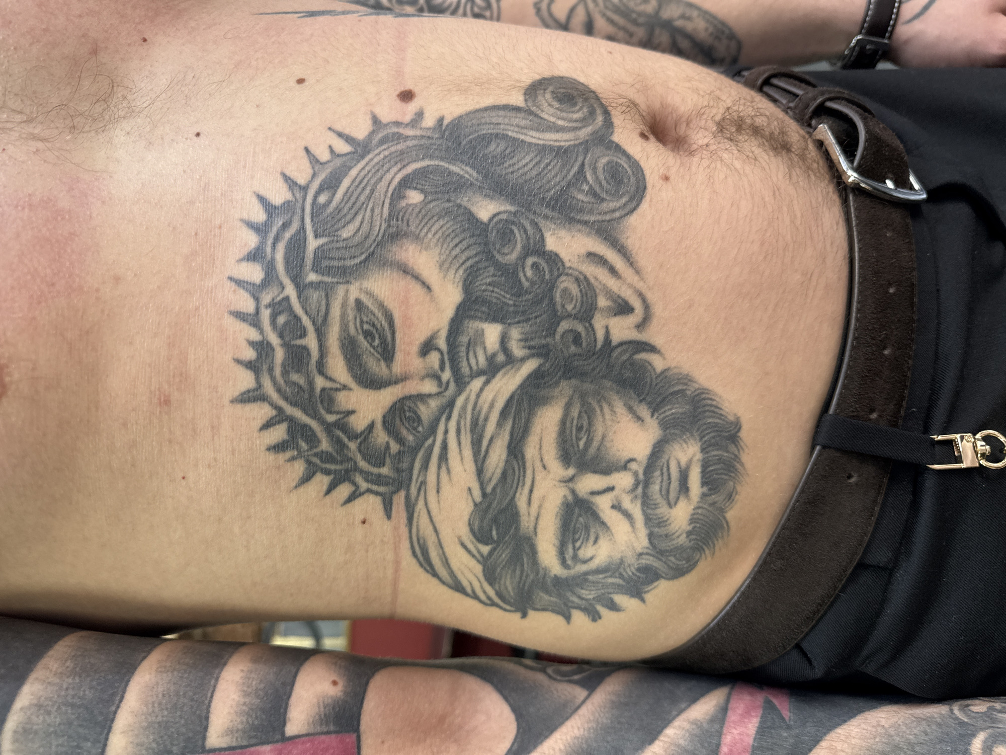 The image is a close-up, angled shot of a person's torso, prominently featuring intricate black and grey tattoos. The central tattoo, located on the right abdomen and ribcage, depicts two intertwined faces. One face, positioned higher, has curly hair and what appears to be a crown of thorns. The second face, situated below and to the right, features a beard and a head covering. Both faces are rendered in a classical or religious portrait style.
The subject's skin shows visible body hair, particularly around the navel and along the waistline, suggesting a male. Several small, dark moles are also scattered across the skin. A dark brown leather belt with a silver-colored buckle is worn around the waist, securing a black garment. In the lower foreground, another tattooed limb—possibly an arm or leg—is visible, displaying bold, dark patterns, including what appear to be stripes or solid black areas, indicative of extensive blackwork.
The scene appears to be an indoor setting, with soft, diffused lighting that emphasizes the details of the tattoos and skin texture. The activity is the display of the individual's body art. There are no discernible location cues within the image itself; however, the context provided states the location is Moscow, Russia. No visible text is present in the media file.