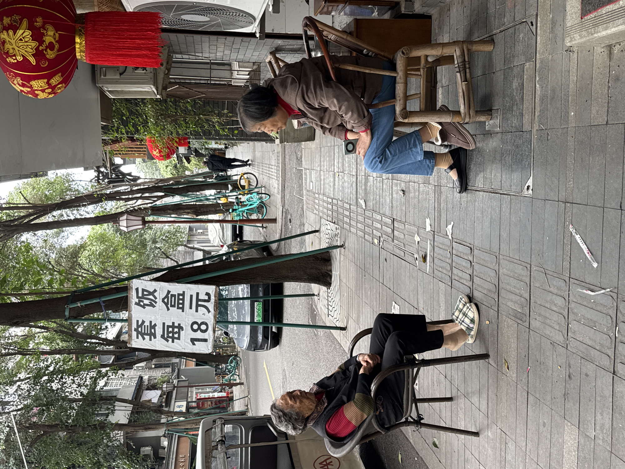 Two elderly women are sitting on chairs on a sidewalk in what appears to be the Qingyang District of Chengdu, China. One woman is seated on a metal-framed chair, wearing black pants, a red shirt, and patterned slippers. Her hands are clasped in her lap, and her head is resting. The other woman is seated on a wooden chair; she is wearing blue jeans, a brown jacket, and closed-toe shoes. She is leaning forward. A red Chinese lantern hangs in the background. A sign with Chinese writing is attached to a tree, and cars and bicycles are visible on the street.