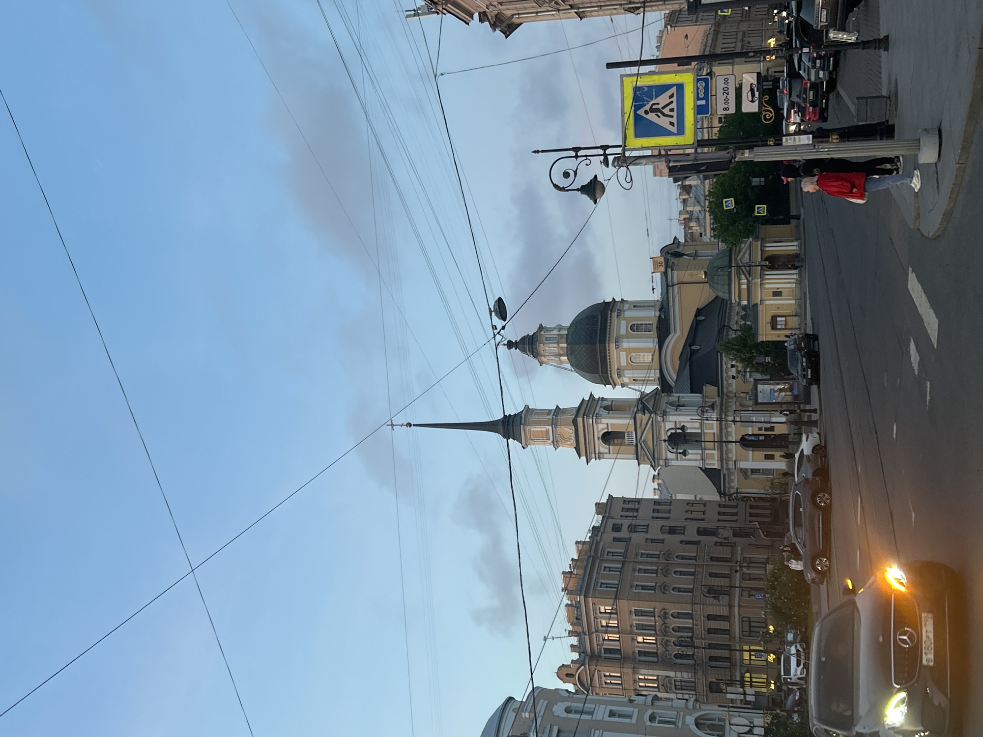The image depicts a street scene in Saint Petersburg, Russia. A church with a green dome and a tall, thin spire stands out against a cloudy sky. Power lines crisscross the sky. The buildings are old and have ornate facades. On the right, a pedestrian crossing sign is visible, along with street lamps. A person in a red jacket is standing near the sign. Cars are parked along the street, including a black Mercedes with its headlights on.