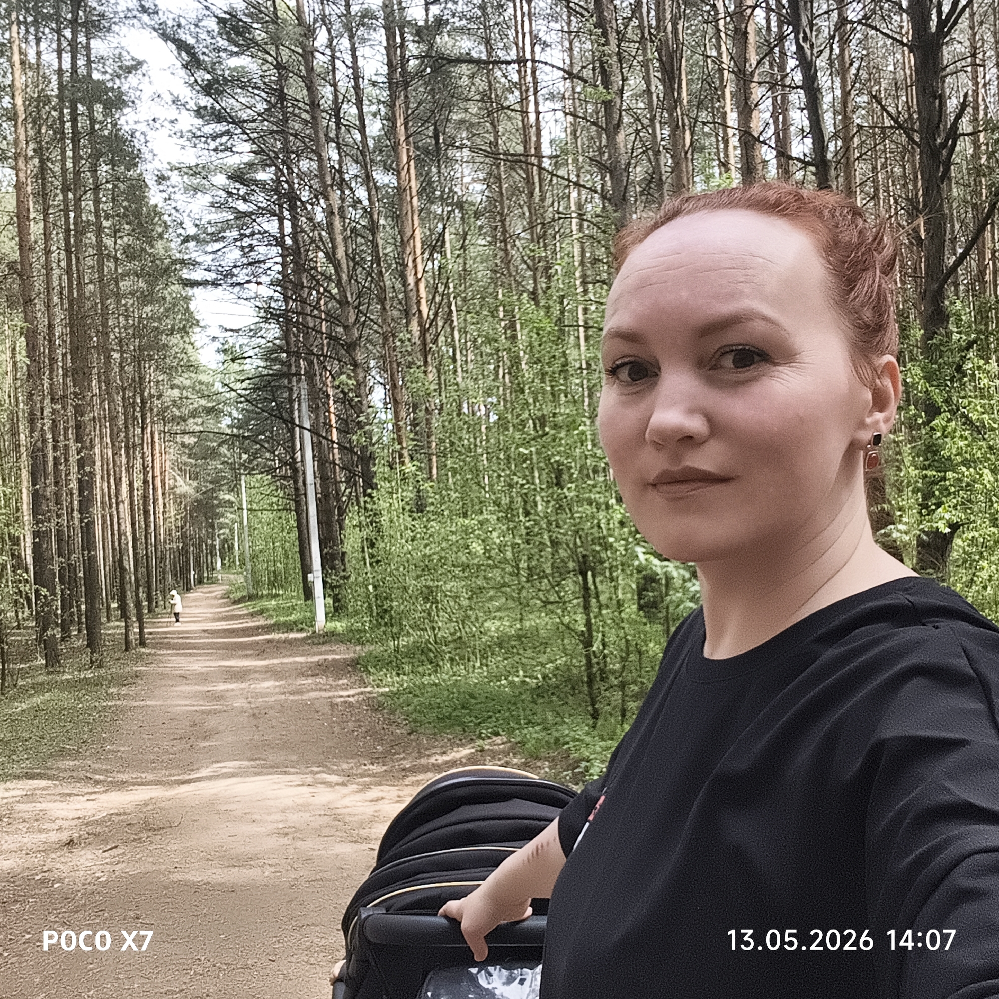 A selfie of a fair-skinned, red-haired woman in a black t-shirt, pushing a black stroller on a dirt path through a dense pine forest. The path stretches into the distance, where a person walks. The surrounding environment consists of tall pine trees and green foliage. The timestamp "13.05.2026 14:07" and the device model "POCO X7" are visible in the lower corners of the image. The location is the city of Chaikovsky, Russia.