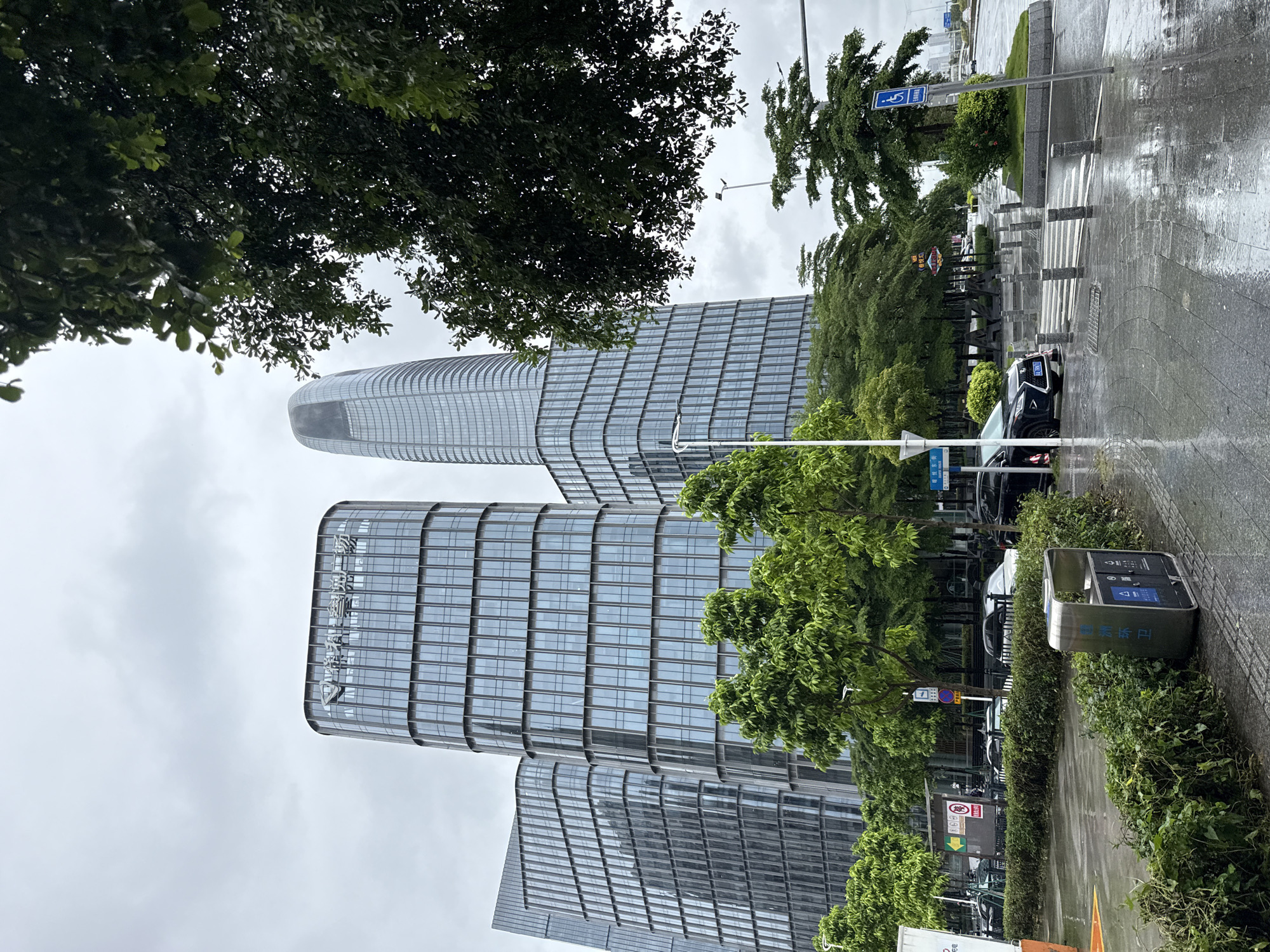 The image depicts a contemporary urban street scene in Haizhu District, China, under an overcast sky. Several modern glass-clad skyscrapers dominate the background, one of which has a distinctive curved facade. Lush green trees line the street and are interspersed between the buildings, some of which are trimmed into hedges. The foreground shows a wet pavement, indicating recent rain, with puddles reflecting the grey sky. A pedestrian crossing with bollards is visible. To the right, a disabled parking sign is prominently displayed. On the left side of the street, a metallic waste bin is situated near a hedge. Cars are parked and driving on the wet road. The overall impression is a well-maintained urban environment, likely a business district, experiencing inclement weather.