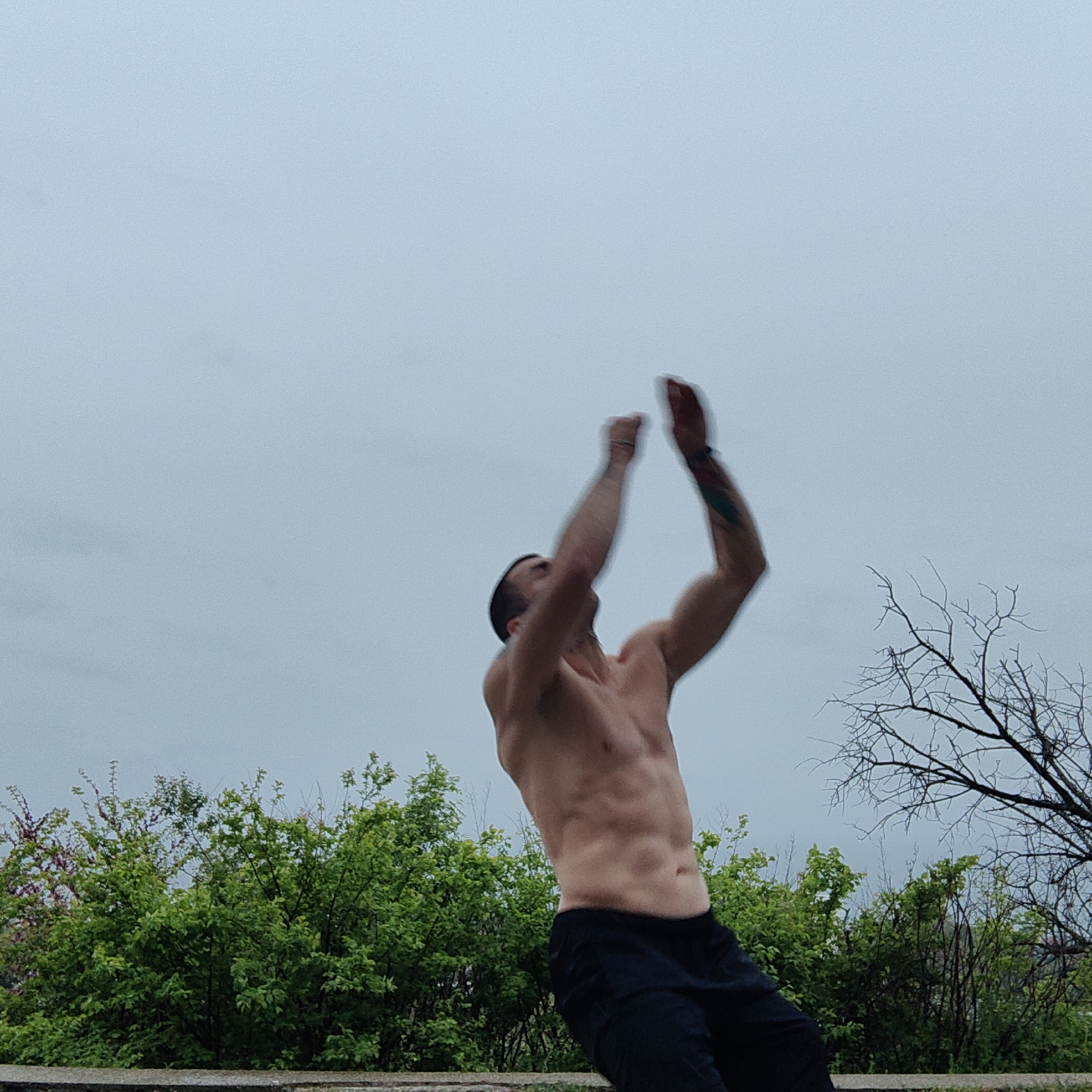 The image displays a shirtless man engaged in a dynamic physical activity outdoors. He is captured mid-action, facing upwards with both arms raised above his head, creating a blur effect due to motion. His head is tilted back, and his gaze is directed skyward. The man exhibits a lean, muscular physique with visible abdominal definition and is wearing dark pants. A tattoo is visible on his right forearm.
The setting features a low, light-colored concrete or stone wall in the foreground. Behind the man, a dense line of vibrant green bushes and leafy trees forms the midground. To the right, a tree with distinct bare, dark branches contrasts with the surrounding foliage. The background consists of an expansive, uniform light blue-grey sky, indicating an overcast day without direct sunlight. The scene suggests an outdoor workout or athletic performance in a natural environment, potentially a park or recreational area in Varna, Bulgaria. No text is visible in the image.