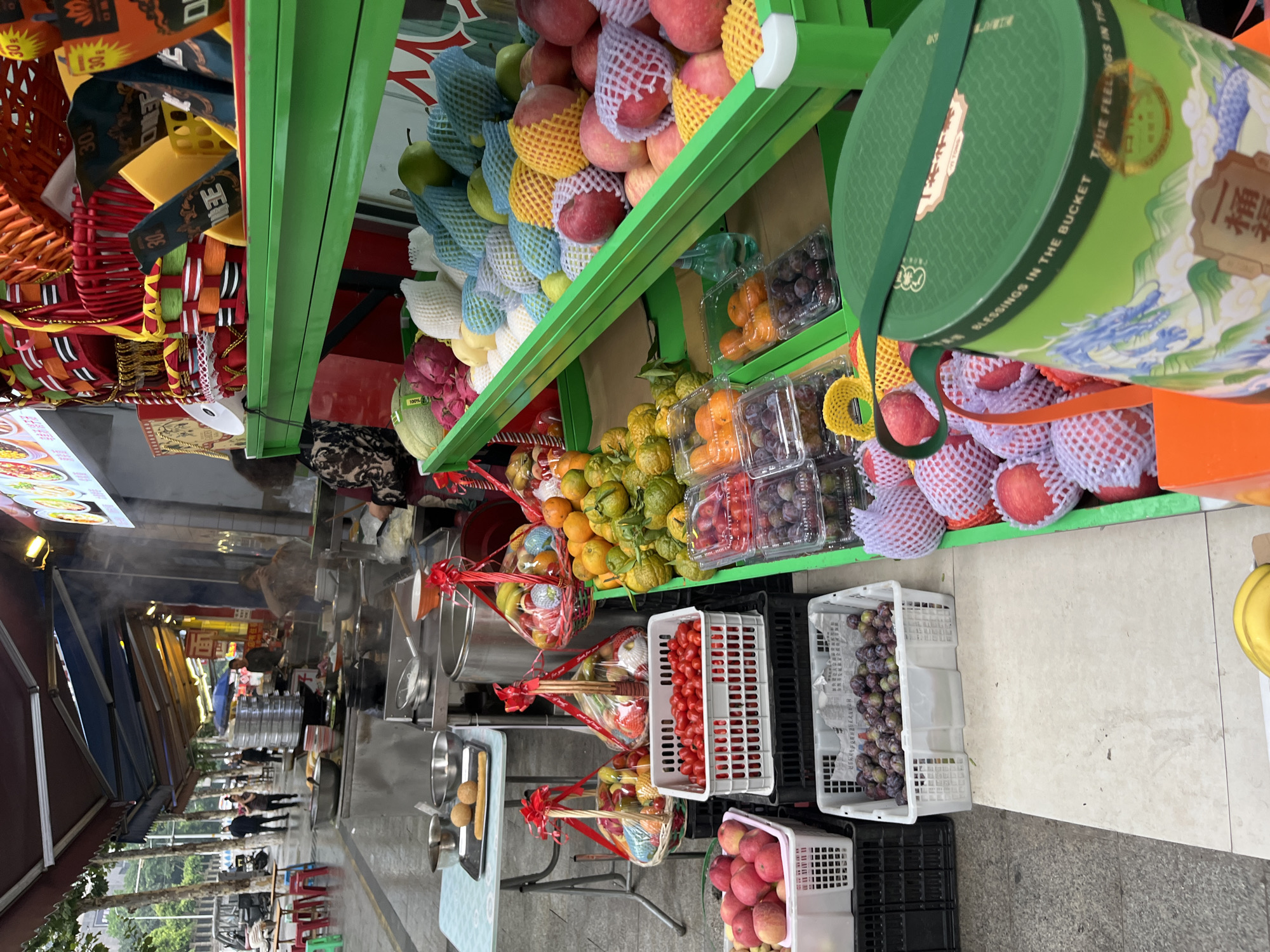 The image captures an outdoor fruit stall in Yuzhong District, China. The stall features a variety of fruits displayed in baskets and crates. Several individuals are visible in the background, some appearing to be vendors or customers. The scene suggests a bustling street market environment, with awnings providing shade and food preparation stations visible.