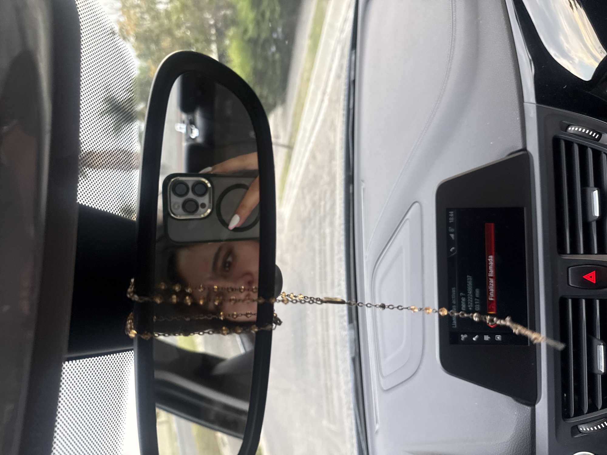 A person is seen in the rearview mirror of a car. They are holding a smartphone, possibly taking a selfie. A beaded necklace hangs from the mirror. The car's dashboard is visible. The car's screen shows a notification in Spanish for a call. The air vents are visible next to the screen. This image was taken in San Bernardino Tlaxcalancingo, Mexico.