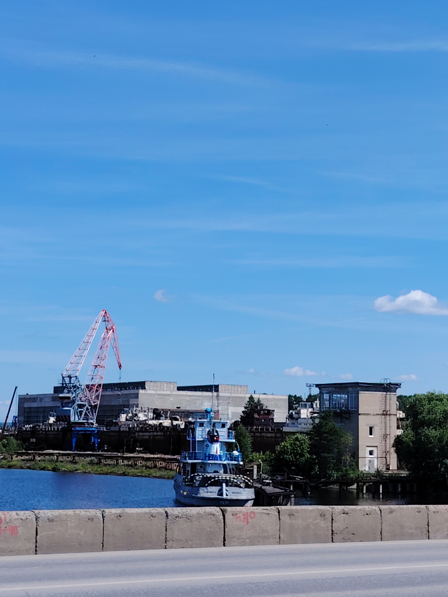 This is a photograph of a river scene in Chaikovsky, Russia, on a clear, sunny day with a bright blue sky. In the foreground, a concrete barrier with visible graffiti runs across the bottom of the frame. Beyond the barrier, a wide, calm blue river occupies the middle ground. On the left side of the river, a large industrial complex is visible, featuring a prominent red and white construction crane reaching high into the sky. Several dark, hulking structures, possibly ships or barges under construction or repair, are docked near the crane. A smaller, blue and white boat is positioned in the water to the right of these industrial elements, appearing to be a tugboat or a small passenger vessel. Further to the right, a tall, light-colored building with a glass-enclosed upper section stands amongst dense green trees. The overall impression is one of a working port or shipyard on a river.