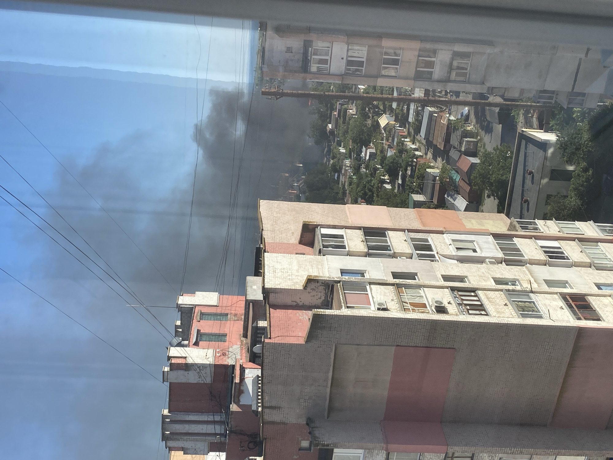 This is an image taken from a window in Chornomorsk, Ukraine, showing a view of an urban area with smoke plumes rising in the distance. In the foreground, there are portions of apartment buildings, characterized by their brick and plaster facades, balconies, and windows. The buildings appear to be multi-story residential structures. Beyond the immediate buildings, a densely packed area of smaller structures, possibly garages or single-story homes, is visible. Trees and vegetation are interspersed throughout this area. A significant amount of dark smoke is billowing into the sky above the distant structures, suggesting a fire or other combustion event. The sky is clear and blue, indicating daytime. Power lines are visible strung across the sky in front of the smoke. The overall scene depicts an active event of smoke emission in an urban residential environment.