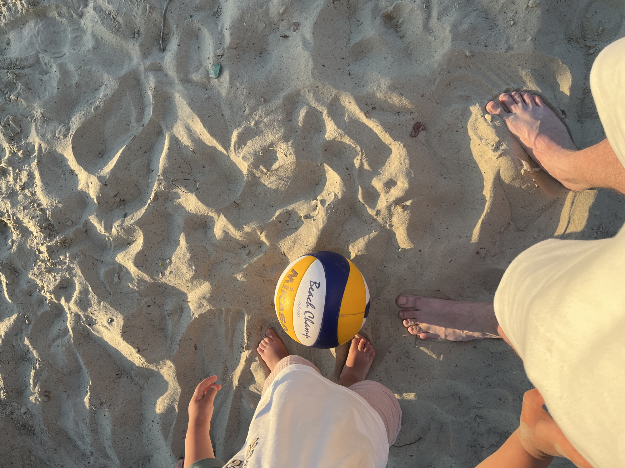 The image shows a close-up, overhead view of several bare feet in the sand, along with a volleyball. The sand is a light tan color and shows the textures of ripples and indentations, with shadows cast by what appears to be late afternoon sunlight.
On the left side of the image, partially visible, are the feet of a child wearing light pink shorts and a white garment. The child's hand is also visible reaching towards the volleyball. Next to the child's feet is a volleyball, predominantly white with yellow and blue panels. Text on the ball is visible, reading "Mikasa" and "Beach Champ VL-300".
In the foreground, a pair of adult bare feet are visible. The feet are positioned as if the person is standing or has just sat down in the sand. The lighting suggests it is either sunrise or sunset.
The scene is a beach. Given that the request specifies Sochi, Russia, this is likely a beach in Sochi, a city on the Black Sea coast known for its beaches and resorts. The sandy environment is consistent with a coastal beach. The presence of a volleyball and bare feet suggests a recreational activity on the beach. There are no buildings or specific city landmarks visible in the immediate frame, but the context implies a public beach area.