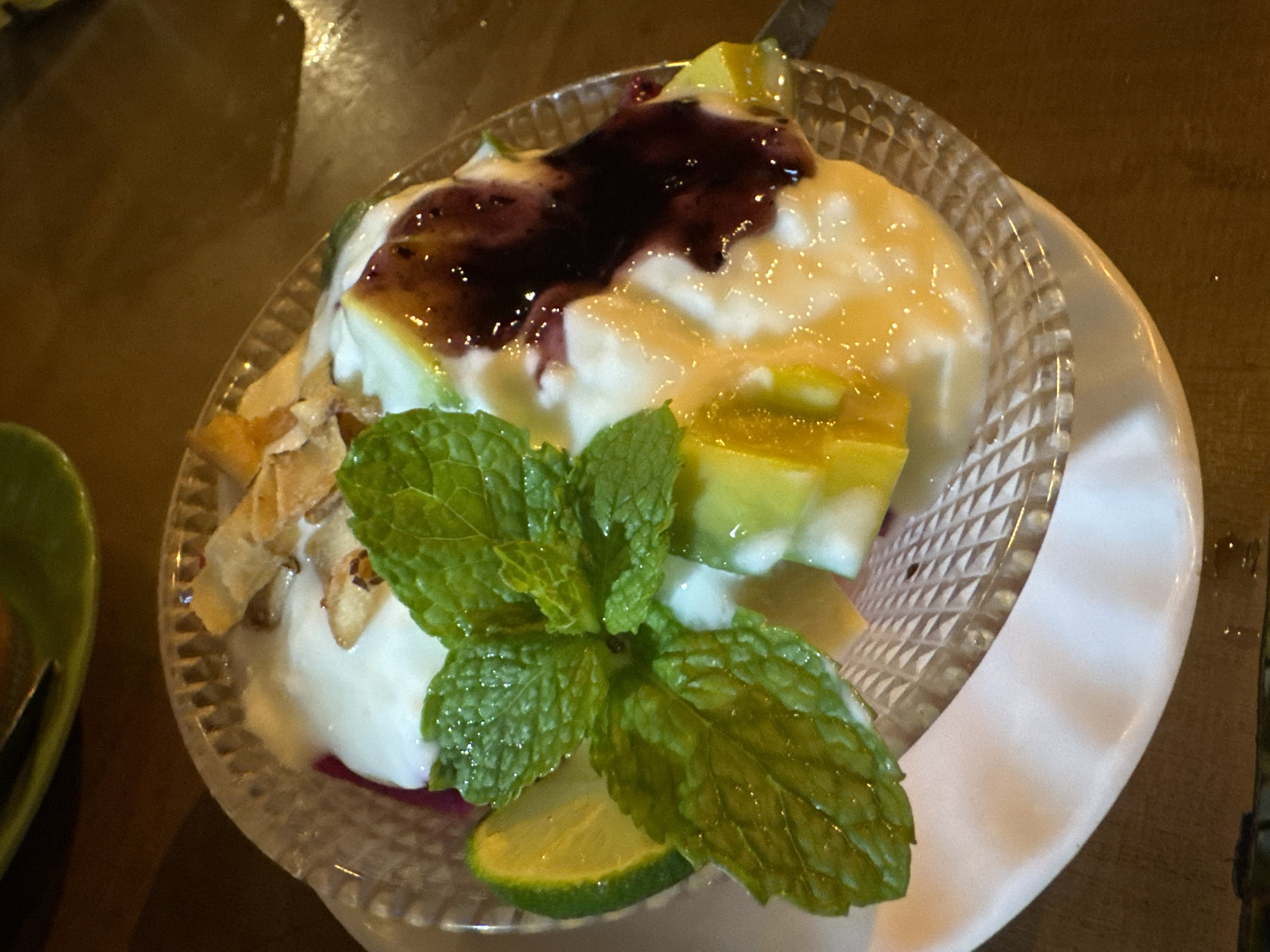 The image presents a close-up, overhead view of a refreshing dessert or breakfast item, likely a yogurt or panna cotta, served in a clear, textured glass bowl resting on a white saucer. The main body of the dish is a creamy white, topped centrally with a glossy, dark purple fruit compote or jam. Surrounding the compote are bright yellow-green fruit cubes, possibly mango or papaya, along with smaller, lighter-colored fruit pieces.
To the left of the dish, a scattering of golden-brown, flaky elements, resembling toasted nuts or coconut shavings, adds texture. A prominent sprig of fresh green mint leaves, dotted with water droplets, acts as a garnish in the foreground, with a thin slice of lime visible beneath it.
The lighting in the photograph is warm and appears to be artificial, suggesting an indoor setting or an evening time of day. The background is soft and out of focus, showing a dark brown surface, likely a wooden table, with a glimpse of another green bowl or plate on the far left. The dish is beautifully presented, indicating it is ready to be consumed. The image captures a culinary detail from Mù Cang Chải, Vietnam. There is no visible text in the image.