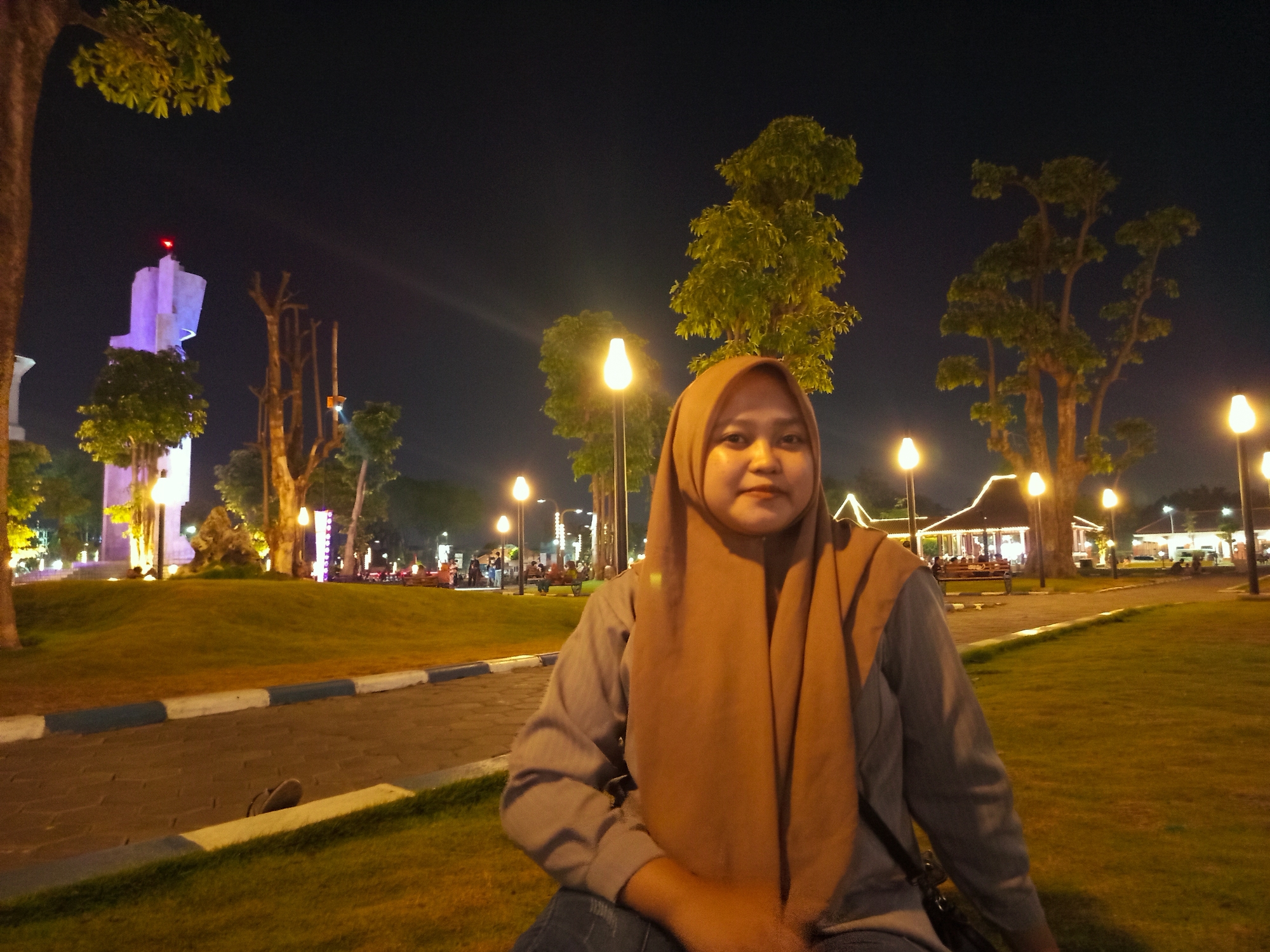 A woman wearing a brown hijab and a grey long-sleeved shirt is sitting on a bench in a park at night, in the city of Sidoarjo, Indonesia. The park is illuminated by streetlights and has several trees and a building in the background. The woman is looking at the camera and smiling. The image is taken from a slightly low angle.