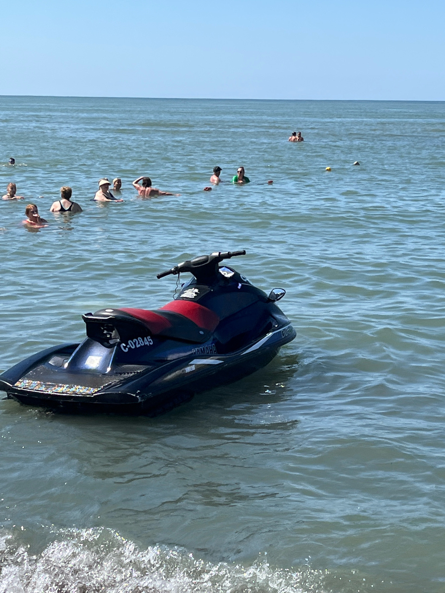 The image depicts a sunny day at a beach in Ureki, Georgia. In the foreground, a dark blue and red jet ski is floating on the water. The jet ski has "C-02845" and "YAMAHA" visible on it. In the background, numerous people are in the water, some swimming or wading, while others are simply enjoying the sea. The water is calm with gentle waves, and the sky is clear blue. The overall scene conveys a leisurely summer day at the beach.