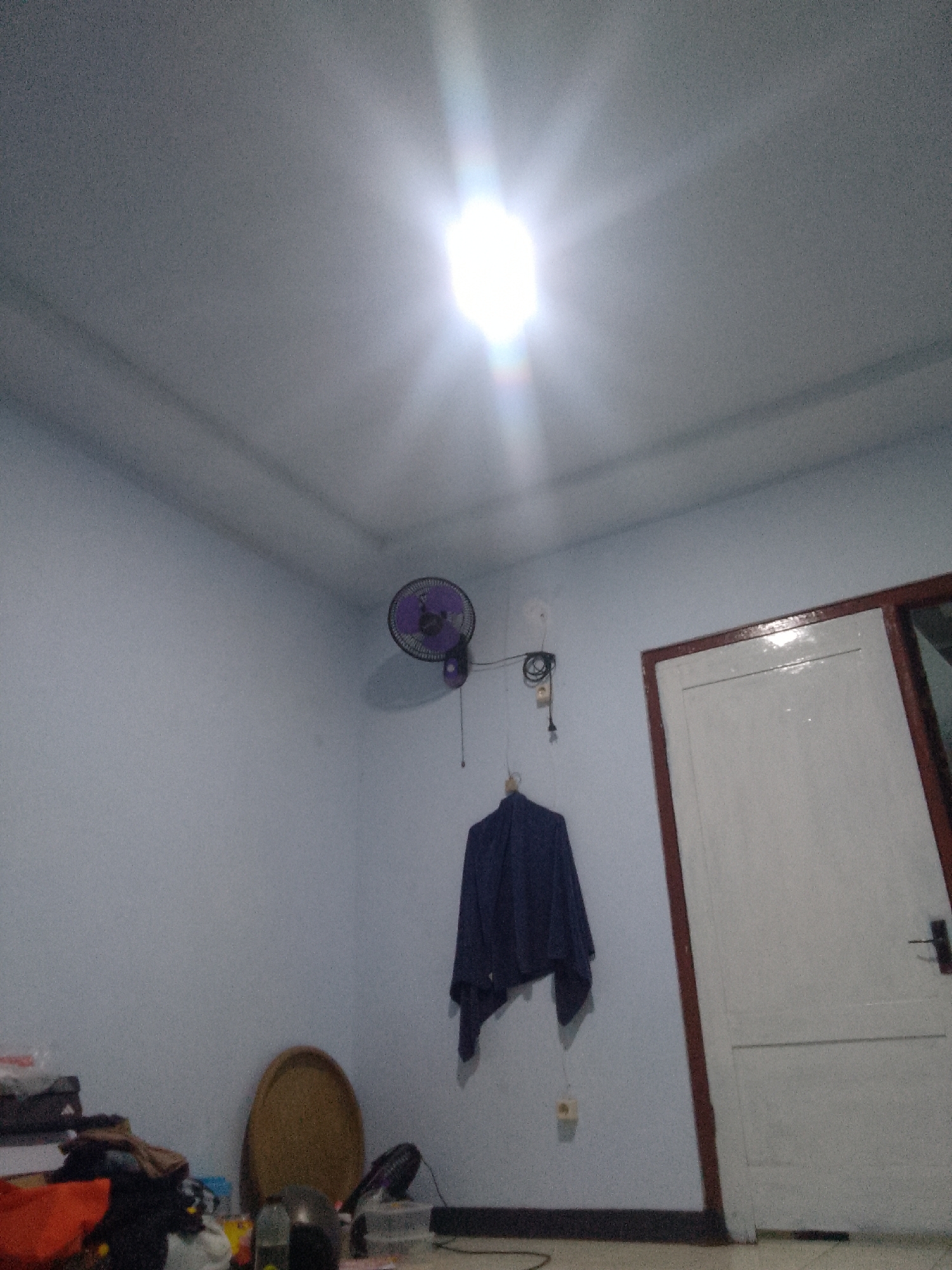 The image depicts an interior scene, likely a bedroom, in Trenggalek, Indonesia. The room is sparsely furnished, with light blue walls and a white ceiling illuminated by a bright overhead light fixture. On the left side of the room, a wall-mounted purple fan is visible, with its cord plugged into a wall socket. Below the fan, a dark blue garment is hanging from a hook. A white door with a dark brown frame is present on the right side of the room. In the lower left corner, a pile of various items, including clothes and bags, is visible. A round, woven mat lies on the floor in the center of the room. The overall impression is of a simple, lived-in space.