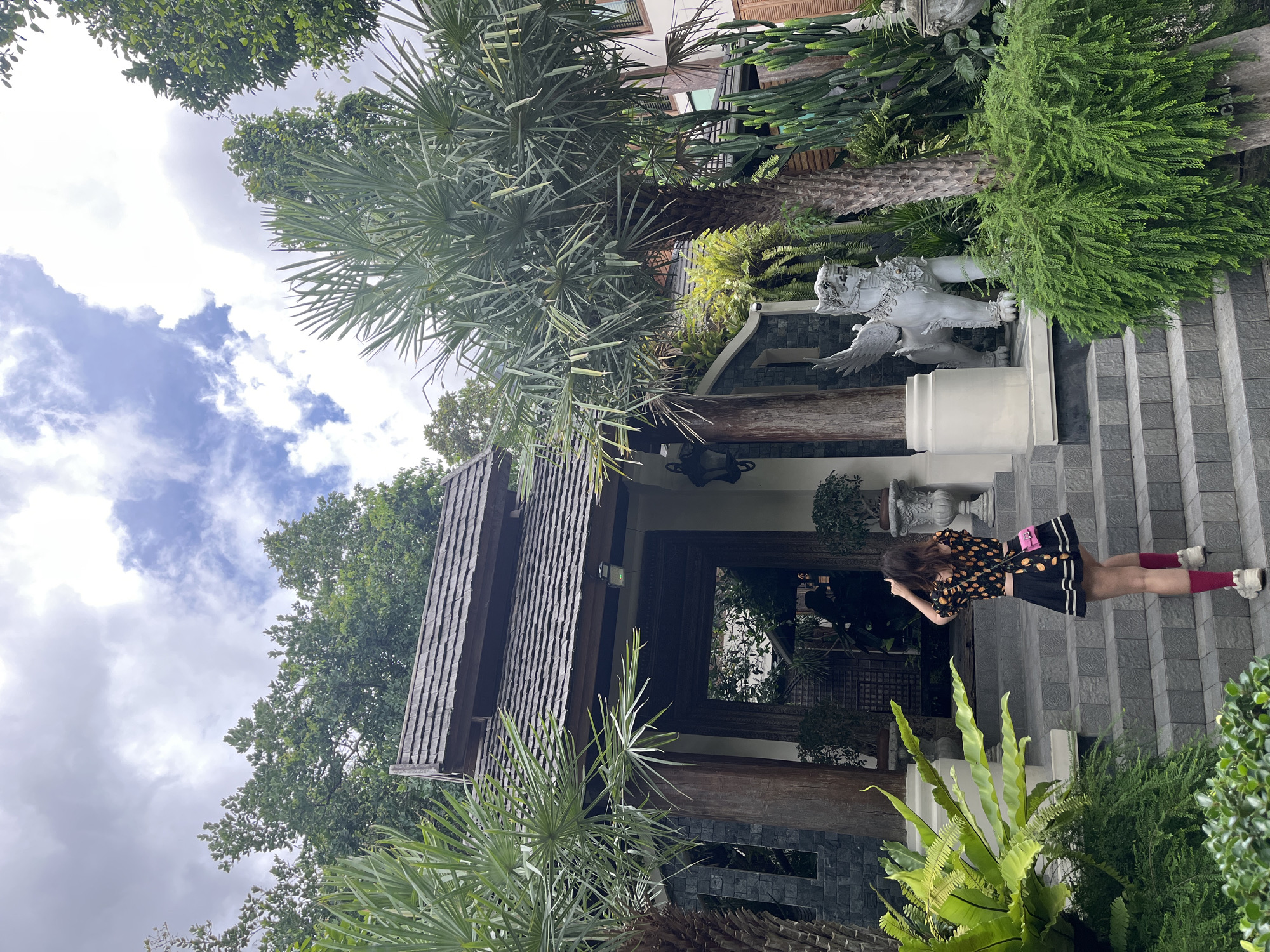 A young woman with dark hair, wearing a black skirt with a yellow and black patterned top and red socks, walks up a grey tiled staircase towards a traditional building with a dark wood entrance. She holds a small pink purse. To her right, a white winged statue of a mythological creature stands near lush green foliage. The building features a dark wood roof with layered shingles and a large, ornate dark wood doorway. The scene is surrounded by dense green trees and plants, including palm trees and cacti. The sky above is cloudy with patches of blue. The image appears to be taken in Suthep, Thailand, given the architectural style and vegetation.