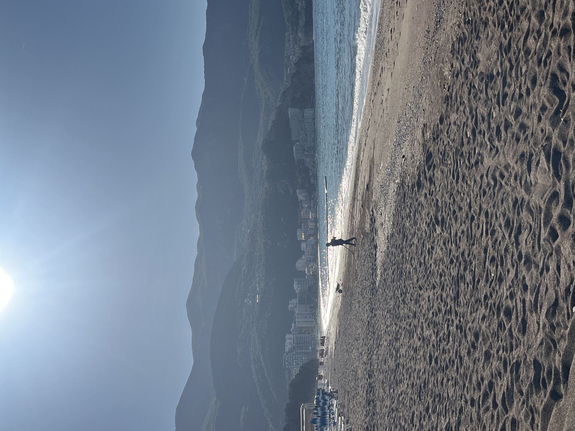 The image depicts a beach scene in Boreti, Montenegro, oriented vertically. A person is walking along the shoreline. The beach has dark sand. Waves are visible near the shore. Behind the beach are buildings and a mountain range. The sun is shining in the upper left corner. Beach umbrellas are visible near the bottom.