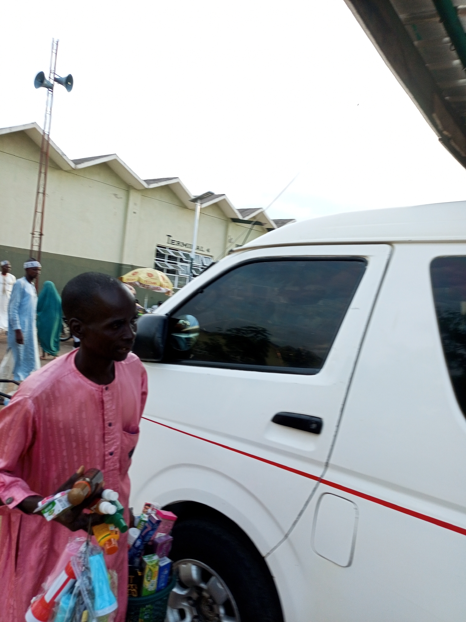The image captures a street scene in Gombe, Nigeria, likely near a transportation terminal. In the foreground, a man wearing a pink traditional outfit stands beside a white minivan. He is holding various items, possibly for sale, including small bottles and colorful packaged goods. A green basket filled with similar items is placed near his feet. Behind him, another man in a blue outfit and a woman in a green hijab are walking. In the background, a building with the sign "TERMINAL 4" is visible, suggesting it's a bus or motor park. A tall pole with two loudspeakers is also present. The overall setting appears to be an open area with a mix of commercial activity and pedestrian traffic.