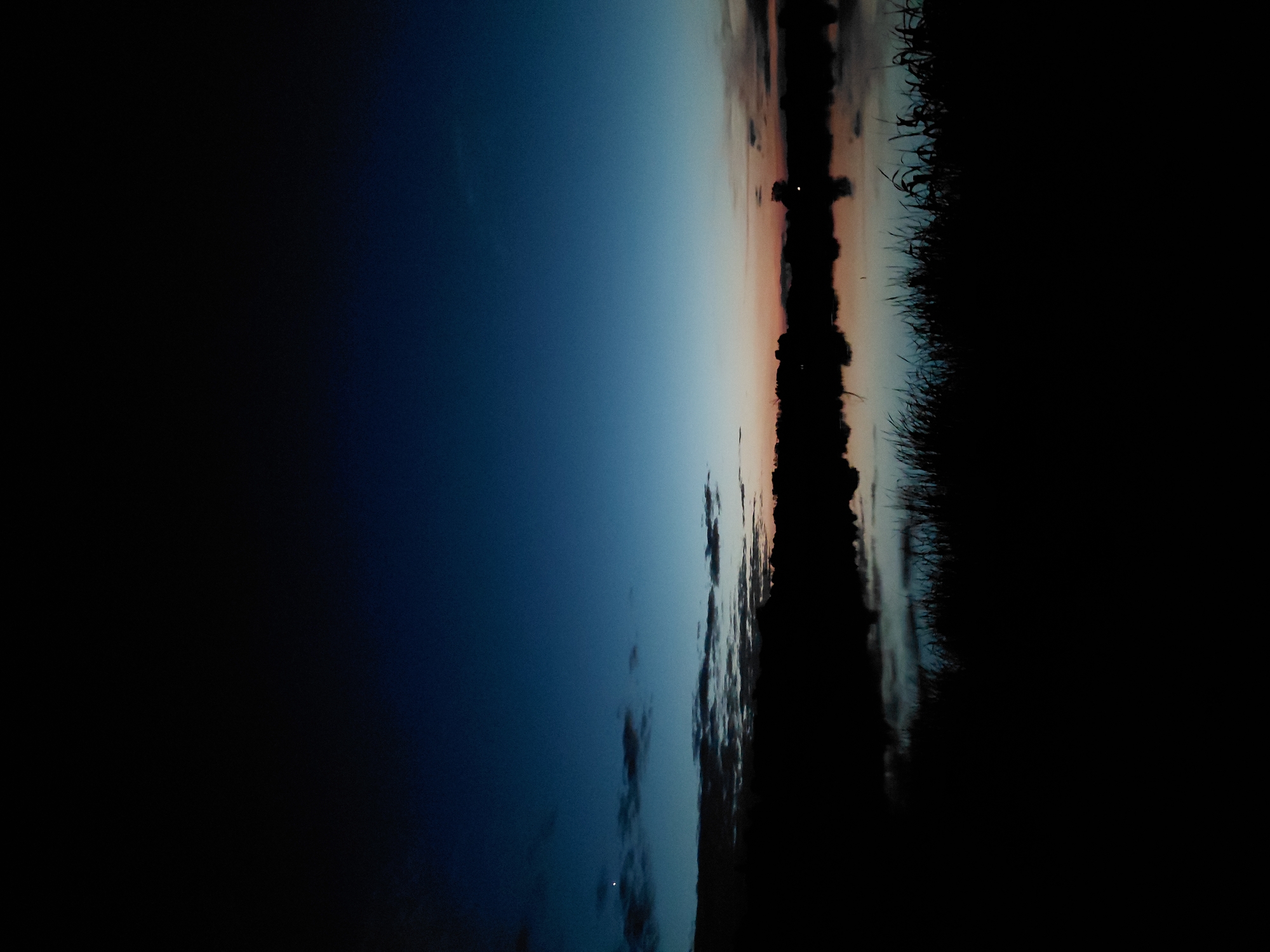 The image depicts a serene twilight scene by a body of water in the city of Ustyvytsia, Ukraine. The sky transitions from a deep blue at the top to lighter hues of blue, pink, and orange near the horizon, indicating the sun has recently set. Silhouetted against this colorful sky is a dark, indistinct line of trees and vegetation along the far bank of the water. The water itself appears calm, reflecting the muted colors of the sky and the dark shapes of the shoreline. In the foreground, tall grasses or reeds are silhouetted, adding to the dark, natural setting. A single bright point, possibly a star or planet, is visible in the upper left portion of the sky. The overall impression is one of tranquility and the quiet beauty of nature at dusk.