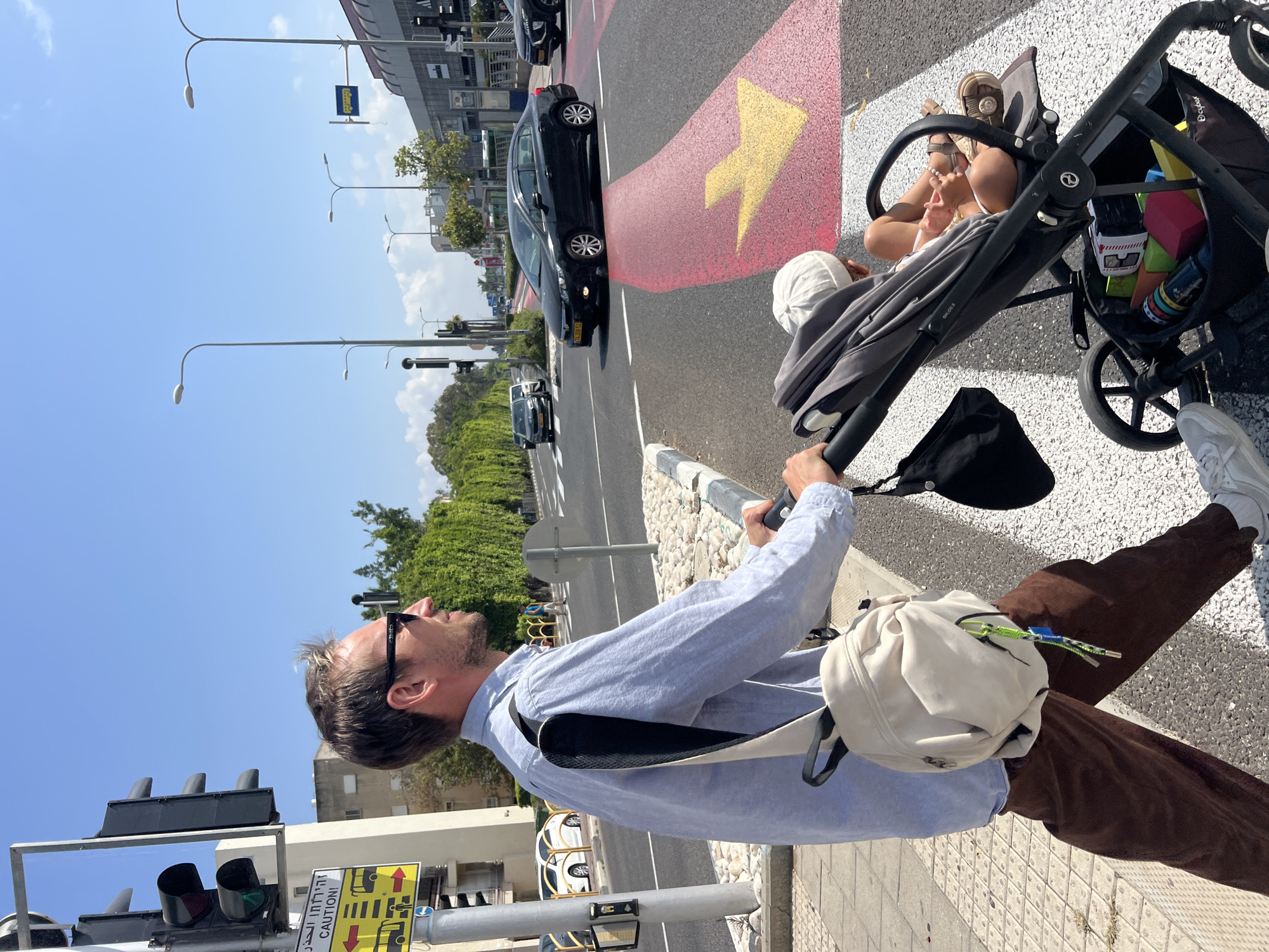 In Kiryat Bialik, Israel, a man wearing sunglasses, a light blue button-down shirt, and brown pants pushes a stroller with a child inside across a pedestrian crossing. The man carries a light-colored bag slung over his shoulder. The stroller has a black frame and a gray fabric seat. The child is wearing a white hat and appears to be looking at toys in the stroller's basket, which contains various colorful items. The pedestrian crossing is marked with white stripes and a large yellow arrow painted on the road. Cars are visible in the background on the street. The sky is clear and blue, indicating sunny weather. Traffic lights and street lamps are also present in the urban environment.