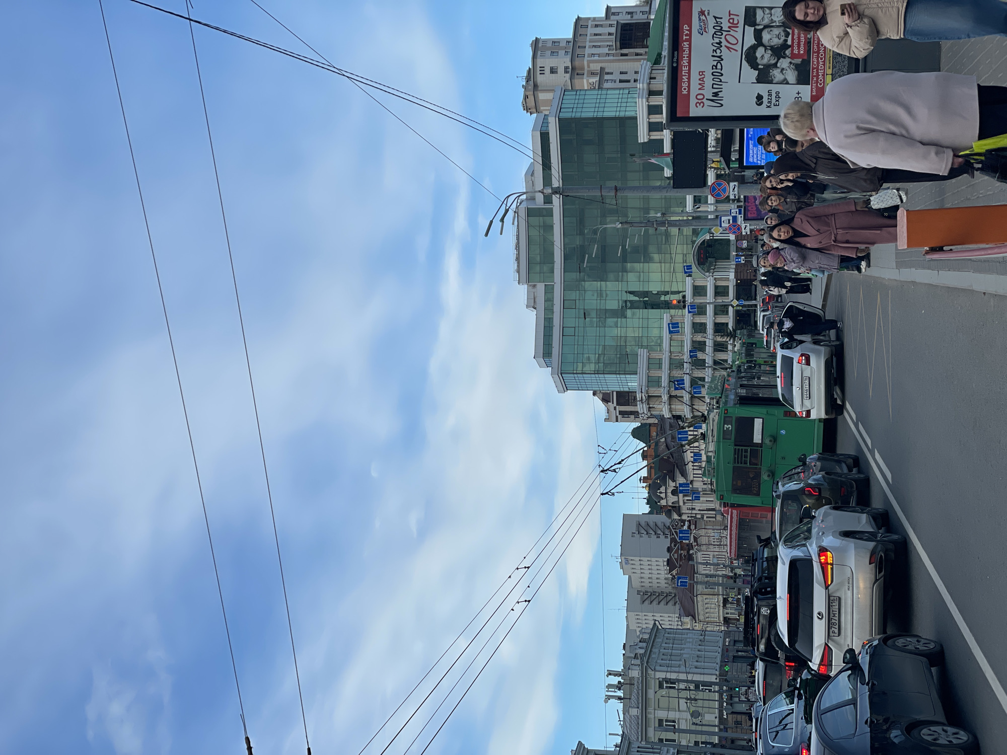 The image captures a vibrant daytime street scene in Kazan, Russia, under a bright blue sky with scattered clouds. The perspective is from the street level, looking down a busy thoroughfare lined with buildings.
On the right side of the image, a sidewalk is populated with numerous pedestrians. A group of people, dressed in various casual outerwear, are seen waiting, some glancing towards the viewer. Among them, a person in a light-colored coat stands prominently with their back to the camera. Behind them, a large billboard displays an advertisement in Russian for an event: "ЮБИЛЕЙНЫЙ ТУР" (Anniversary Tour), "30 МАЯ" (May 30), "Импровизаторы" (Improvisers), "10 лет" (10 years). It also mentions "Kazan Expo," "3+", "дополни КОНЦЕРТ" (additional concert), and "БИЛЕТЫ НА САЙТЕ ОРГАНИ" (tickets on the organizer's website), ending with "COMEDYCONCE."
The multi-lane road in the foreground and center is experiencing a traffic jam, with various vehicles bumper-to-bumper. A silver Lexus RX 300 SUV is clearly visible with the license plate P787MT 116. Another white car has the license plate 8446TT 716. Further down the road, a green bus is part of the queue. Overhead, numerous black power lines crisscross the sky, suggesting the presence of electric public transport infrastructure.
The buildings framing the street present a mix of architectural styles. On the right, modern high-rises with reflective glass facades dominate the skyline, contrasting with older, more traditional structures visible in the background. Traffic lights and street signs are also discernible, contributing to the urban environment. The scene depicts a typical busy day in the city, with people going about their routines amidst traffic.