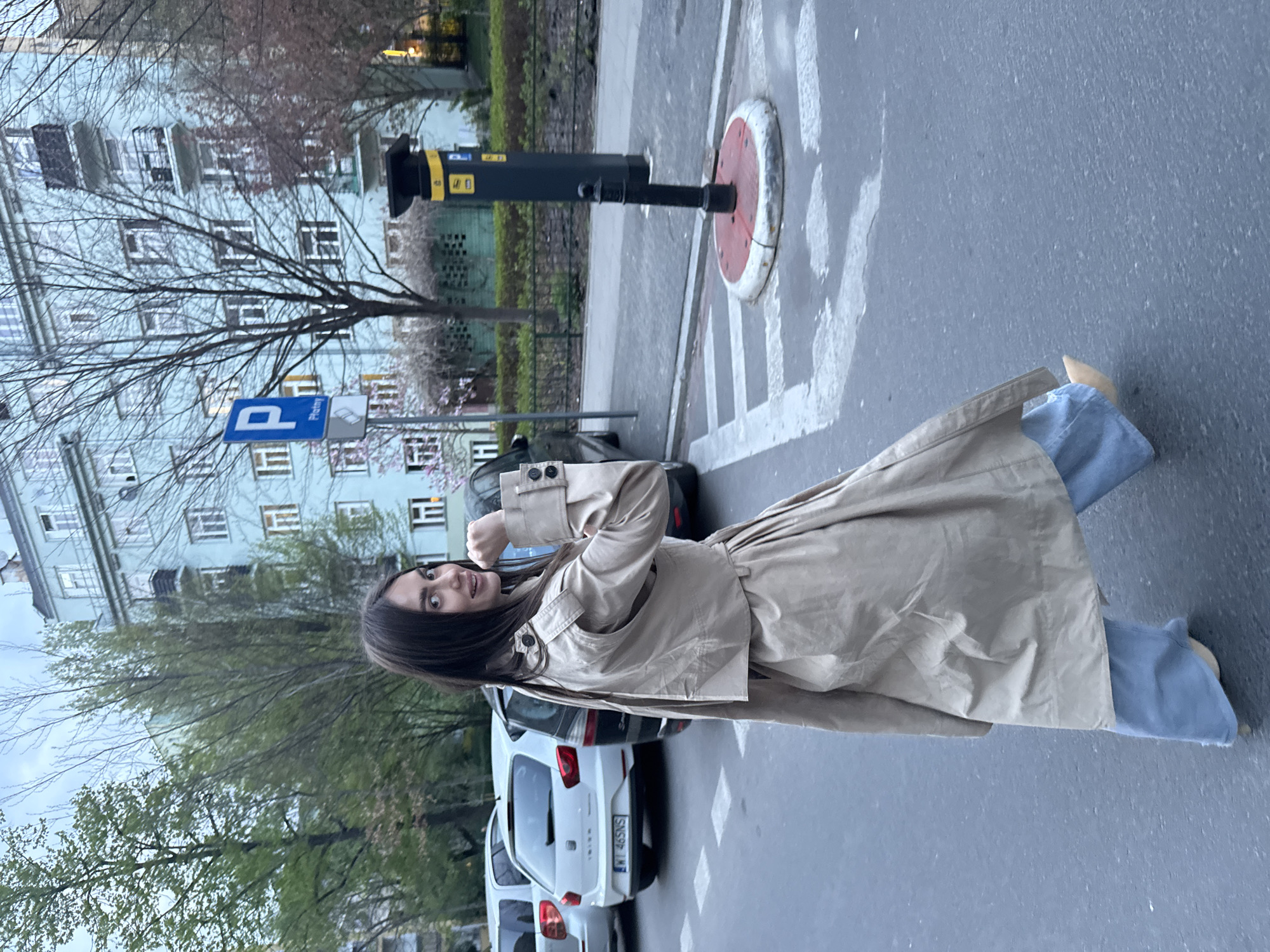 A woman with long dark hair stands on a gray paved street, wearing a light brown trench coat and light blue jeans, with a building in the background. The building is tall, light blue, and has many windows. A blue parking sign is visible on the left, along with trees that indicate a park or green area. Several cars are parked parallel to the street. The license plate of a white car reads "WI 46SNS." There's a traffic safety bollard in the midground. The scene appears to be in Warsaw, Poland.