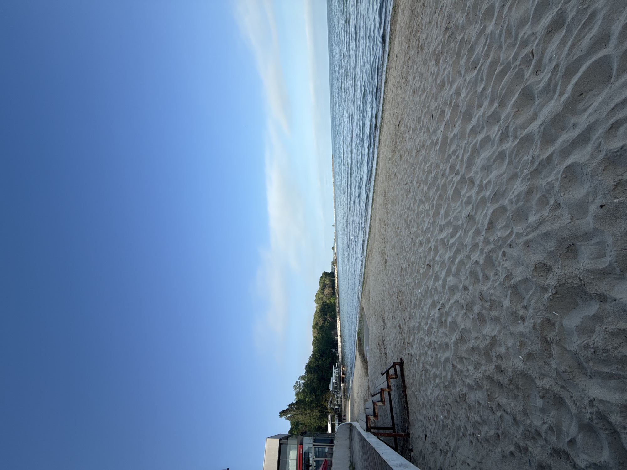 The image depicts a beach scene in Varna, Bulgaria. The beach is sandy, with patterns of wind and footprints visible. The Black Sea lies adjacent to the sand. A line of trees and buildings separate the beach from the city. Stairs of metal are at the border of the beach. The sky is blue with some clouds. In the lower-left corner of the image, there is the top of building with a glimpse of its red interior.