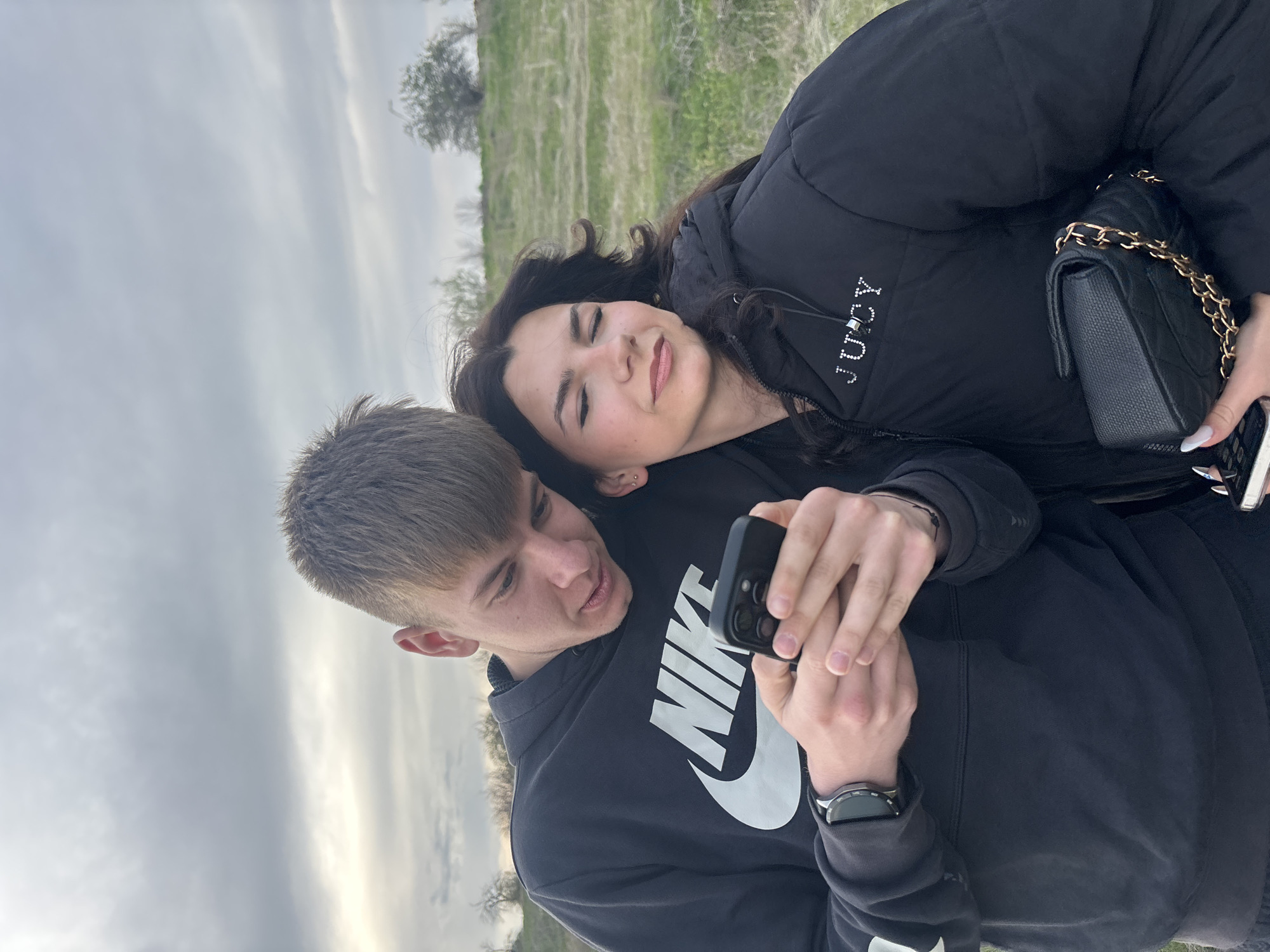 A young man and woman are lying together outdoors, with the man holding a smartphone. The man wears a dark Nike hoodie and a wristwatch. The woman is nestled against him, wearing a dark puffy jacket with the word "Juicy" on it and holding a dark, quilted purse with a gold chain. The background features a field or meadow and a cloudy sky. The location is not discernible as Tsar Asen, Bulgaria.
